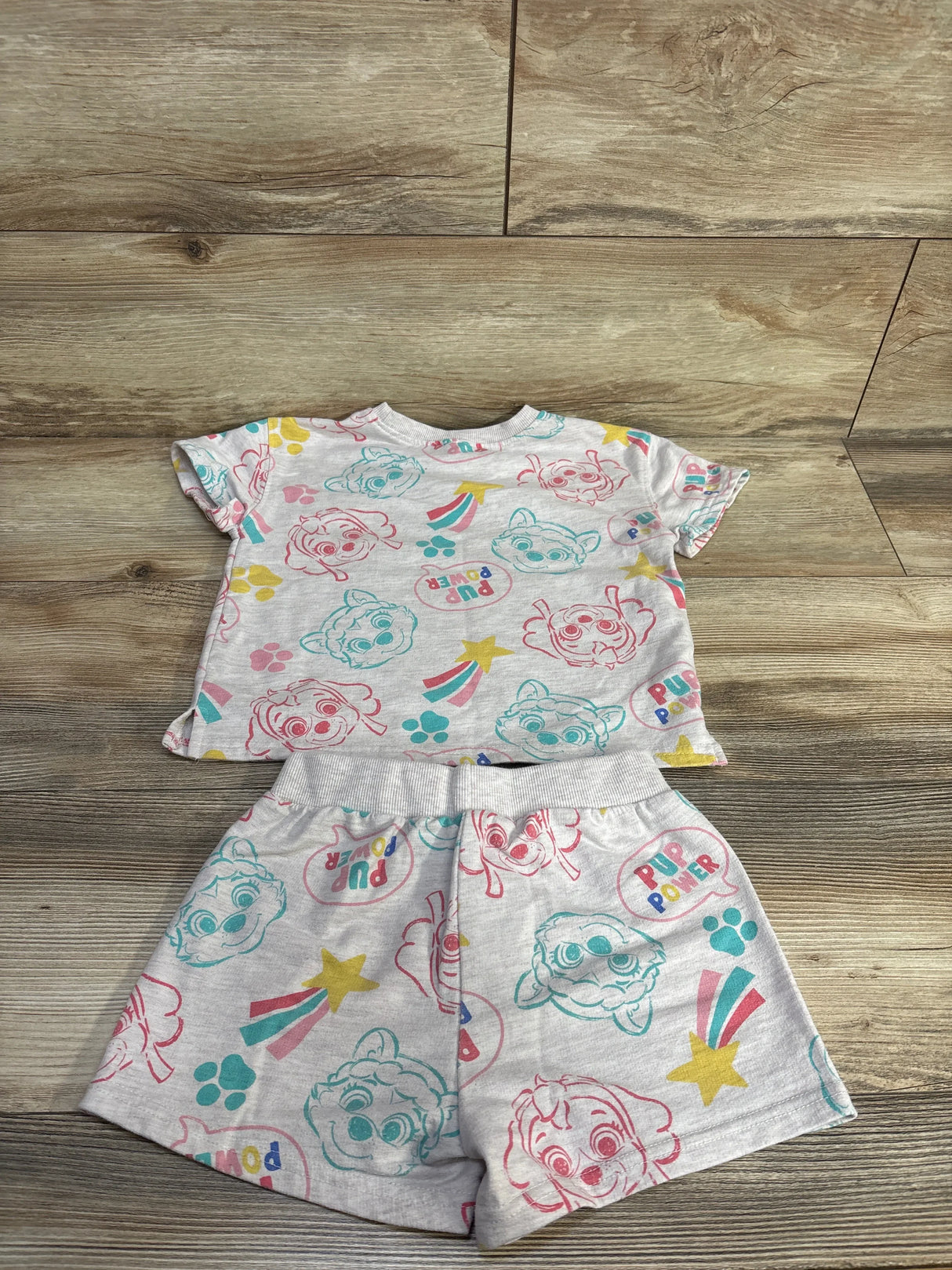 Flat lay of grey PAW Patrol two-piece shirt and shorts showing multicolor character and paw-print pattern on wood background