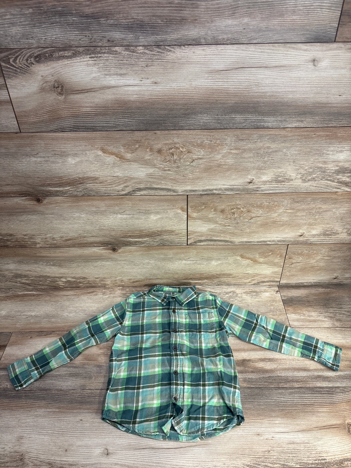 Green and blue plaid long-sleeve button-up shirt laid flat on wooden floor background