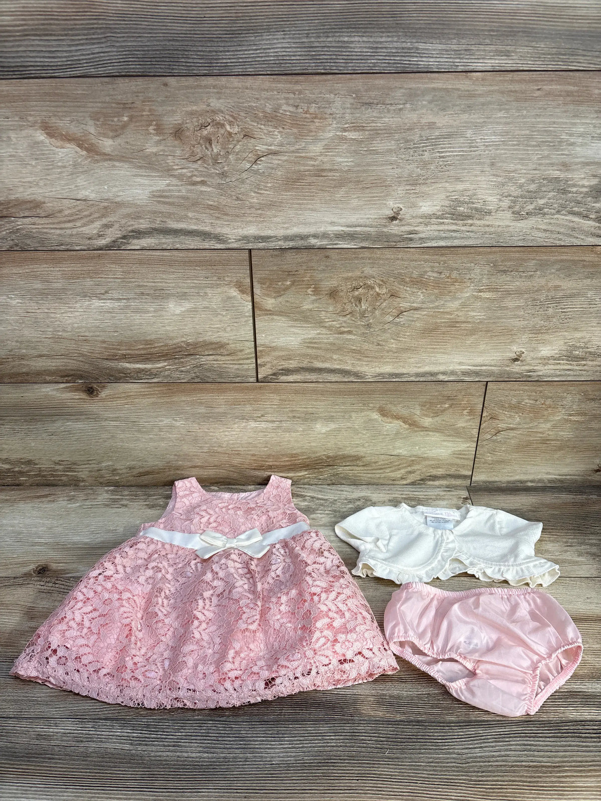 Front view of Bonnie Baby three-piece lace dress set showcasing pink lace dress, bloomers, and white cardigan on wood-look background