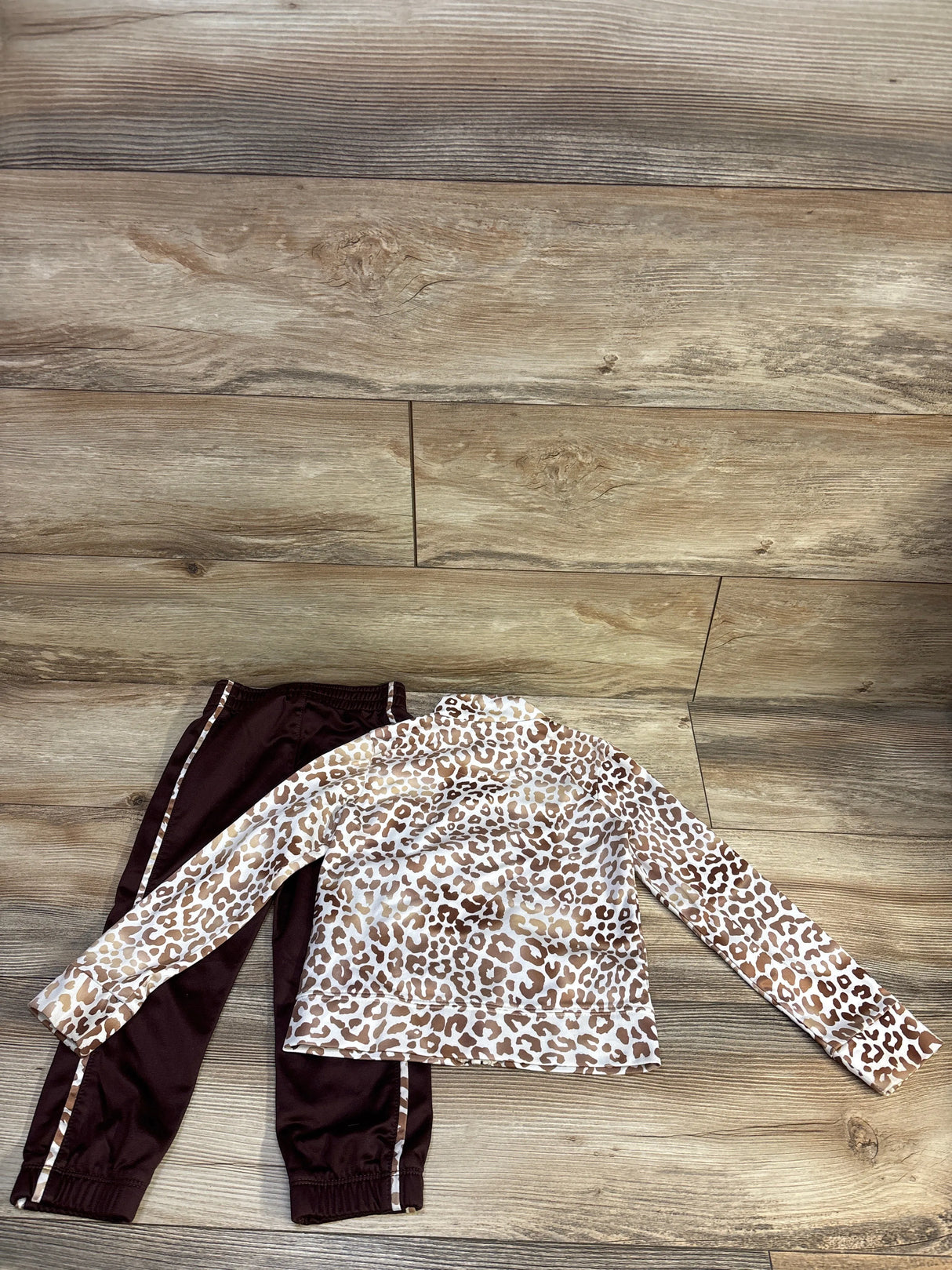 Flat lay of Nike toddler 2-piece leopard print tracksuit showing beige leopard long-sleeve top and dark brown jogger pants with matching leopard side stripes on wood floor