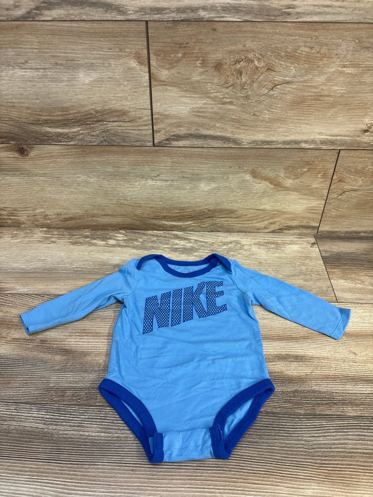 Flat-lay front of light blue Nike long-sleeve 6M bodysuit showing dotted NIKE logo and dark blue trim on wood background