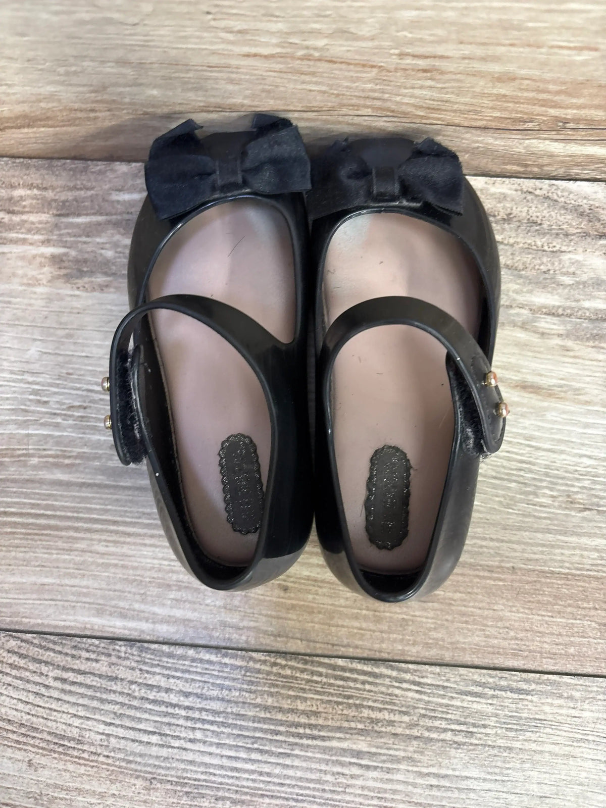 Top view of Mini Melissa Ultragirl Bow jelly flats in black, size 8, showing bow detail and Mary Jane strap on wood floor