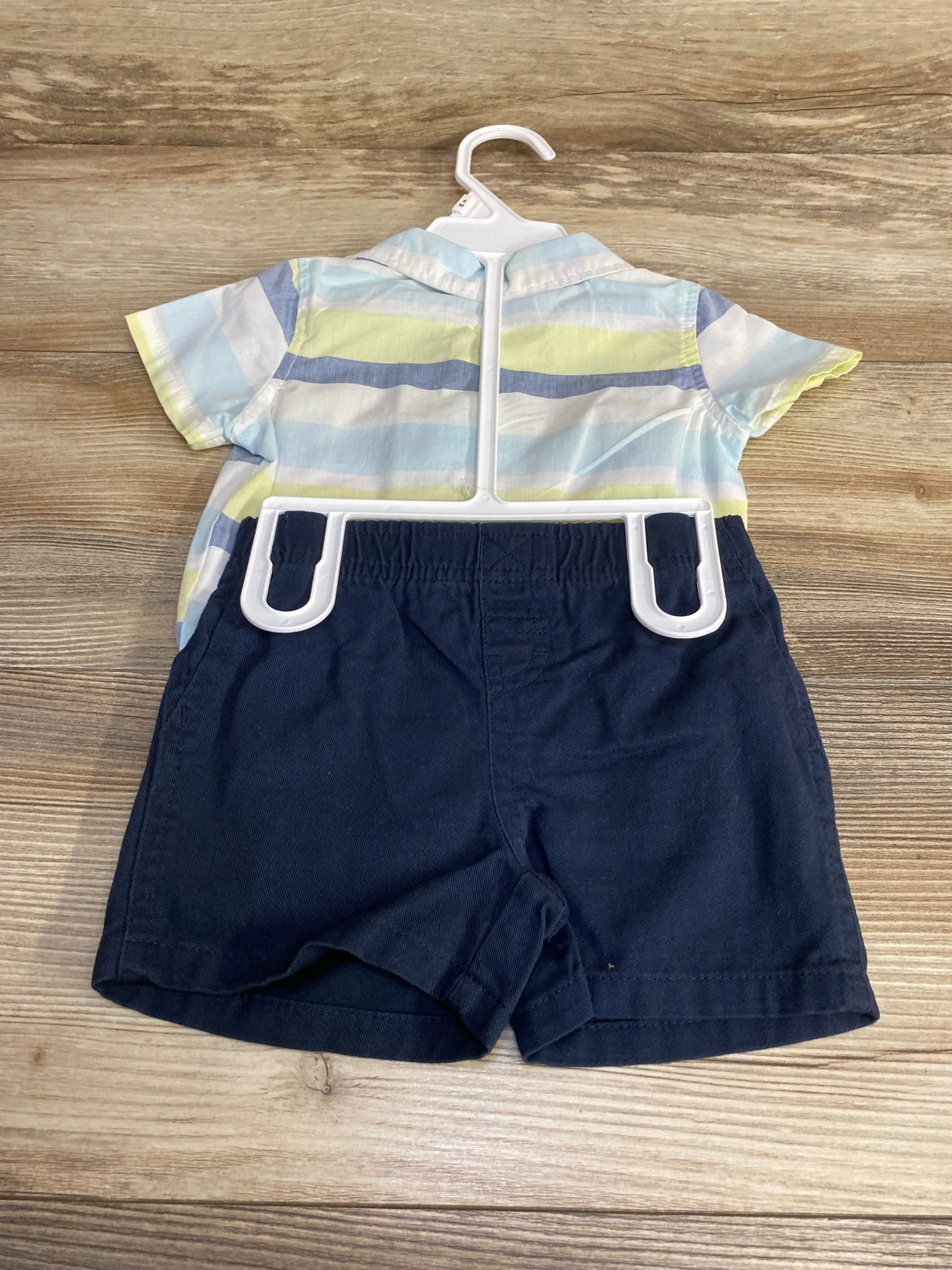 Back view of 2-piece striped baby bodysuit and navy shorts size 3-6M on hanger on wood floor showing pastel stripes and elastic waistband