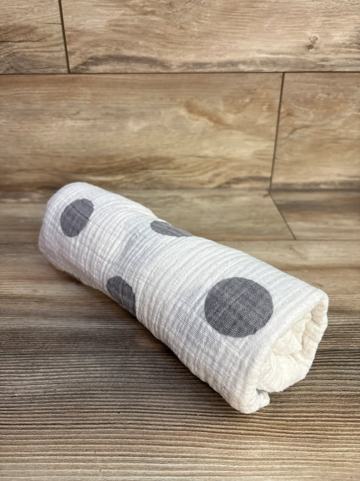 Rolled view of Momo Bebe muslin swaddle blanket showcasing white fabric with gray polka dots on a wood floor