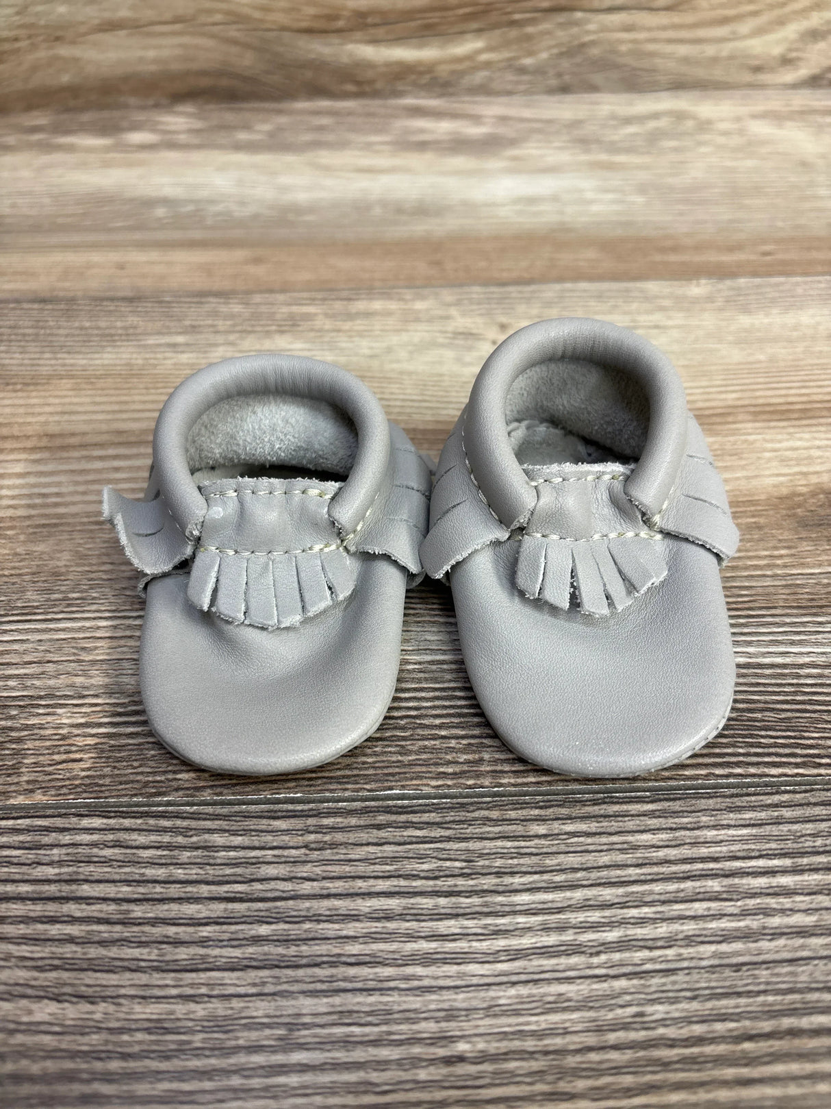 Pair of soft gray leather baby moccasins with fringe detail on wooden surface