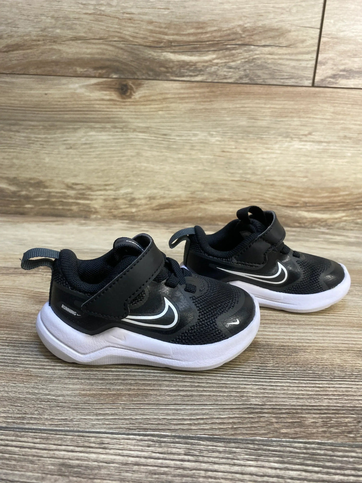 Side view of Nike Cosmic Runner TD toddler sneakers in black and white showcasing Velcro strap and cushioned white sole on wood floor