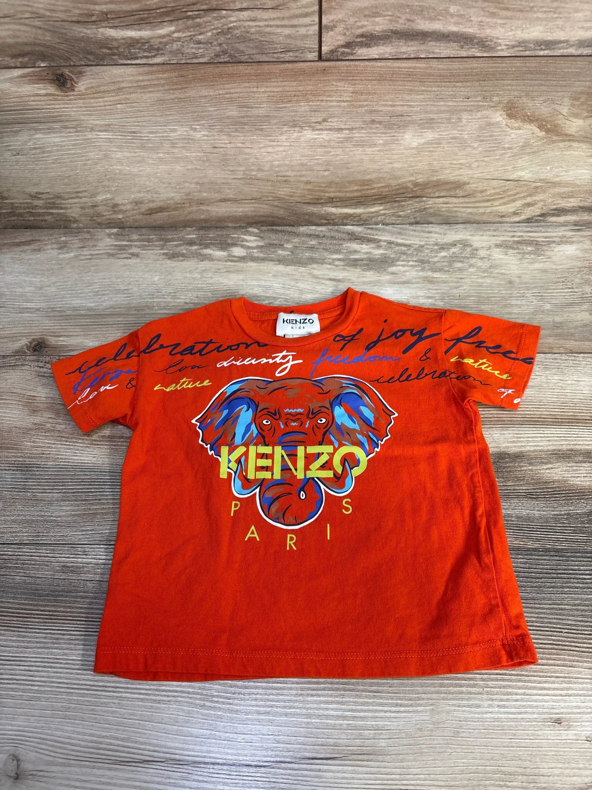 Flat lay of orange Kenzo Kids toddler T-shirt showing multicolor elephant graphic, yellow KENZO PARIS text on wood floor