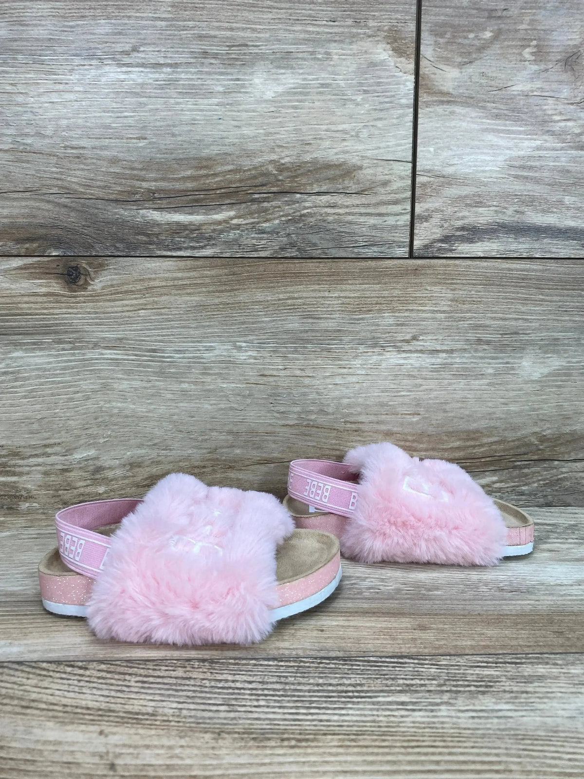 Angled view of toddler pink faux fur sandals with fluffy strap, elastic back band and glitter midsole on wood background