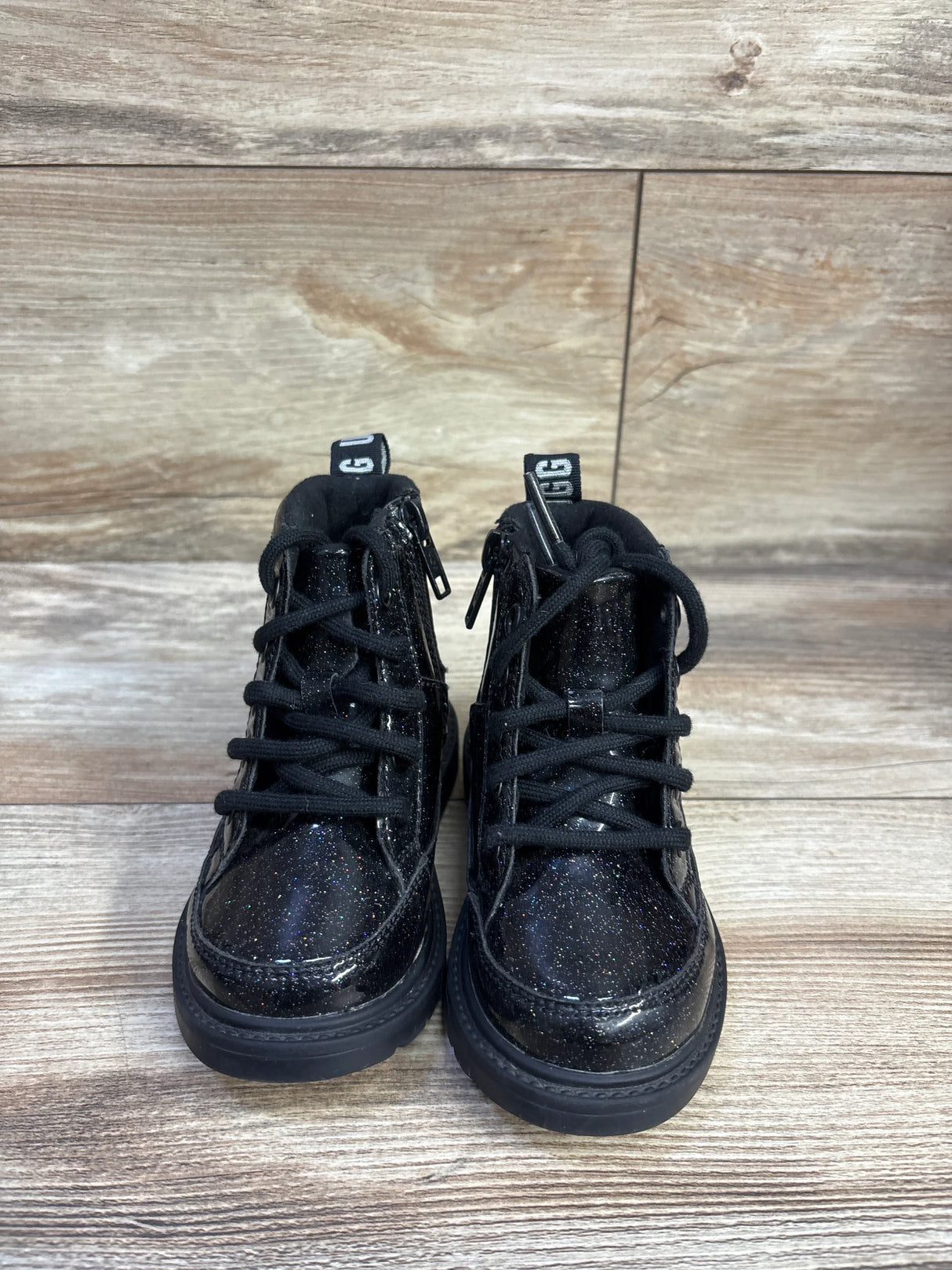 Front view of UGG Kids Robley glitter boots showing black glitter upper, laces and side zippers on light wood floor background