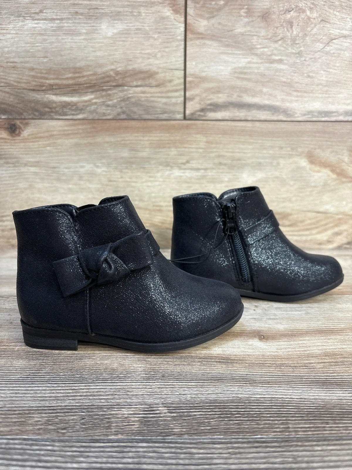 Black toddler ankle boots with side zipper and bow, sparkly finish, on wood background