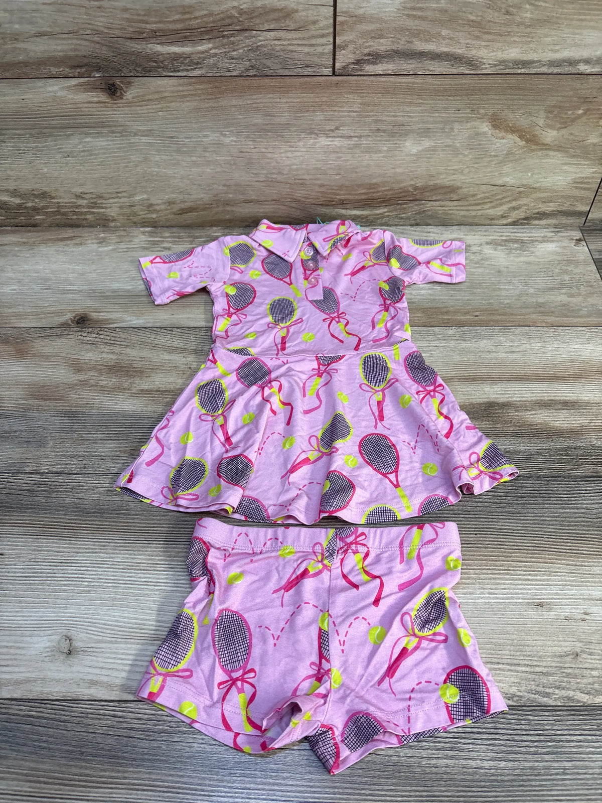 Front view of pink Gigi & Max Serena tennis polo dress and matching shorts set showcasing tennis racket print on wood background