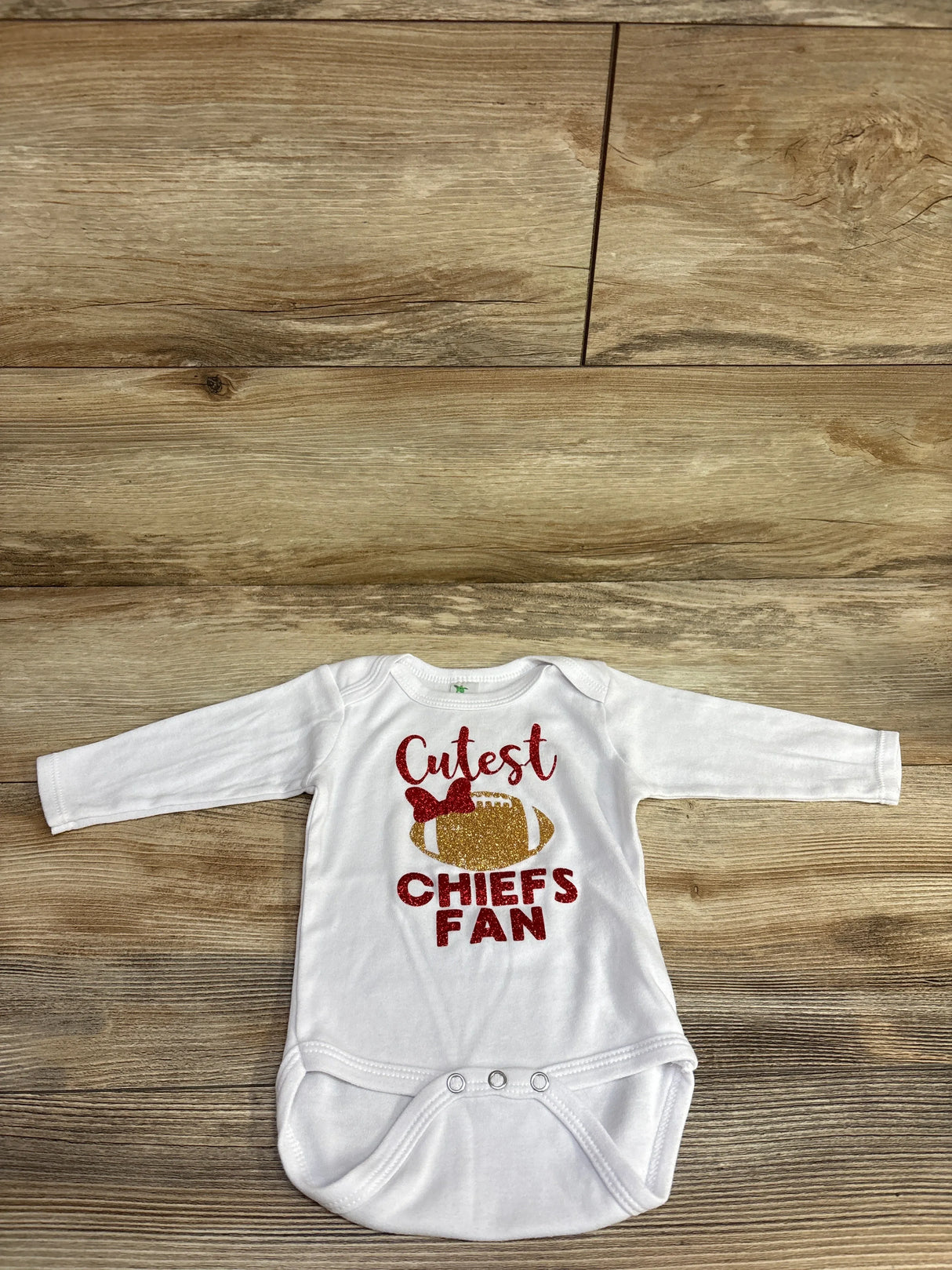 White baby onesie with glitter text saying Cutest Chiefs Fan and a football with a bow on wooden background
