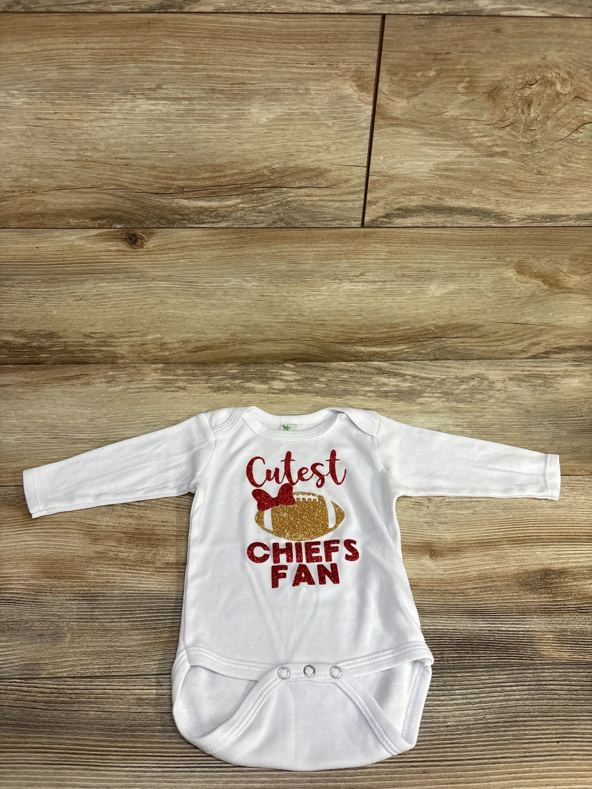 White baby onesie with glitter text saying Cutest Chiefs Fan and a football with a bow on wooden background