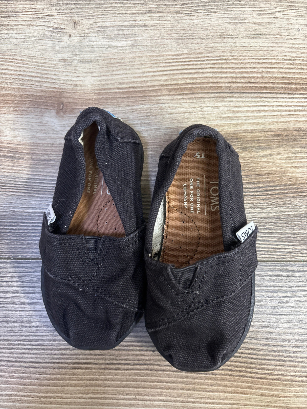 Top-down view of TOMS Tiny Alpargata baby shoes showcasing black canvas upper and TOMS side tag with wood plank background