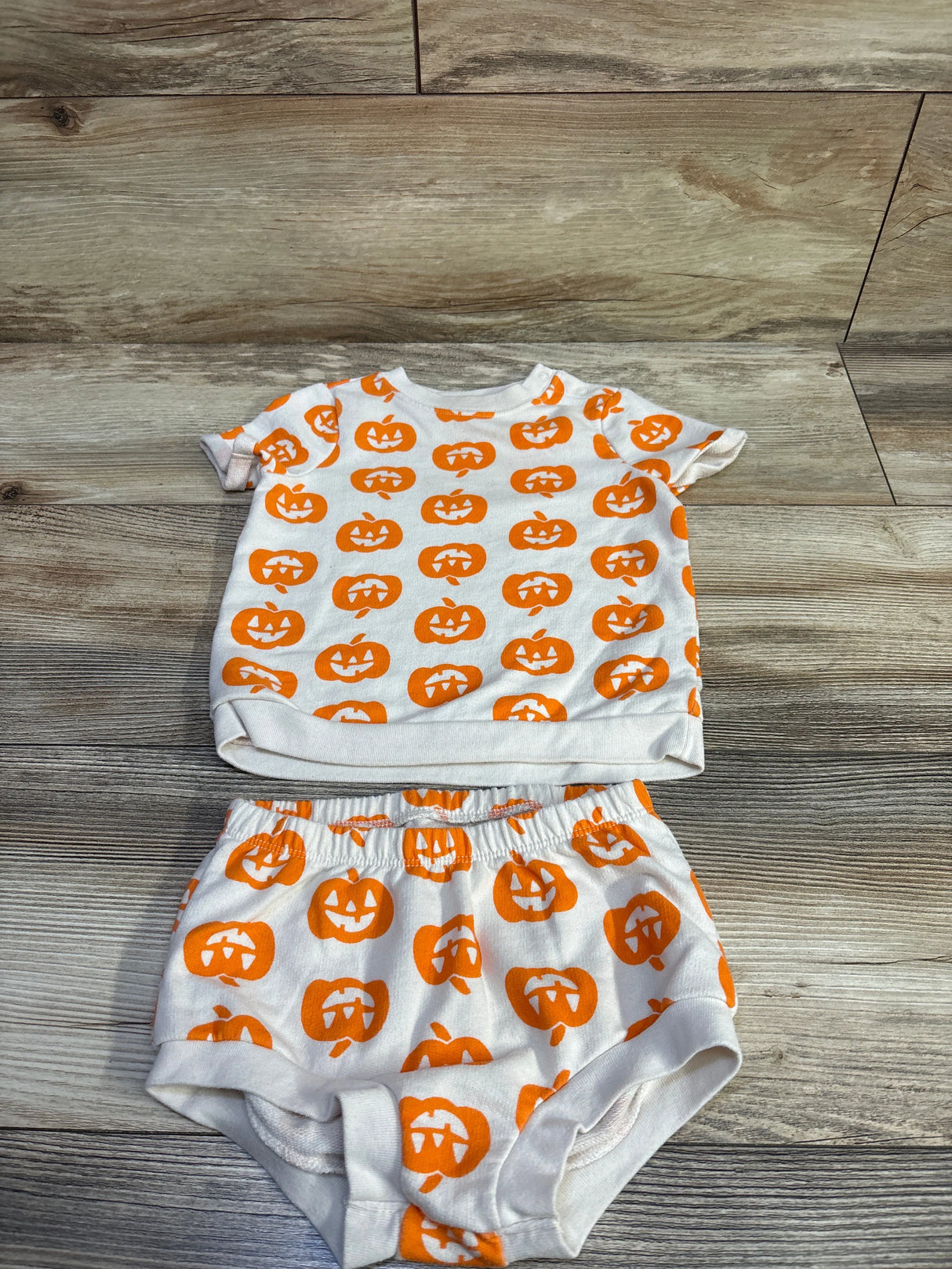 Flat lay of Cat and Jack 2 piece ivory toddler shirt and bloomers set showcasing orange jack-o’-lantern print on wood floor background