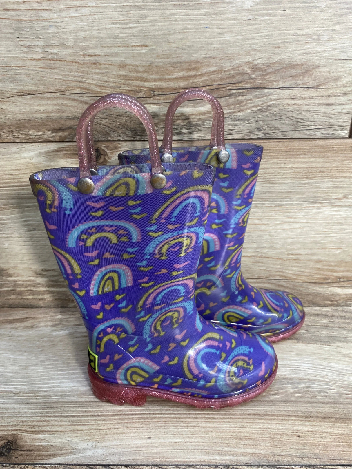 Purple kids rain boots with rainbow and heart pattern, pink glitter handles, wood background