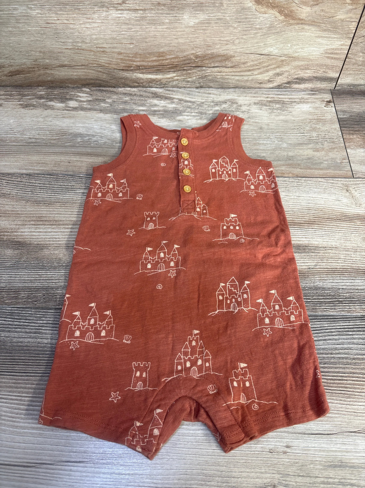 Flat lay of rust brown Little Co sandcastle romper with white print and wooden front buttons on light wood floor