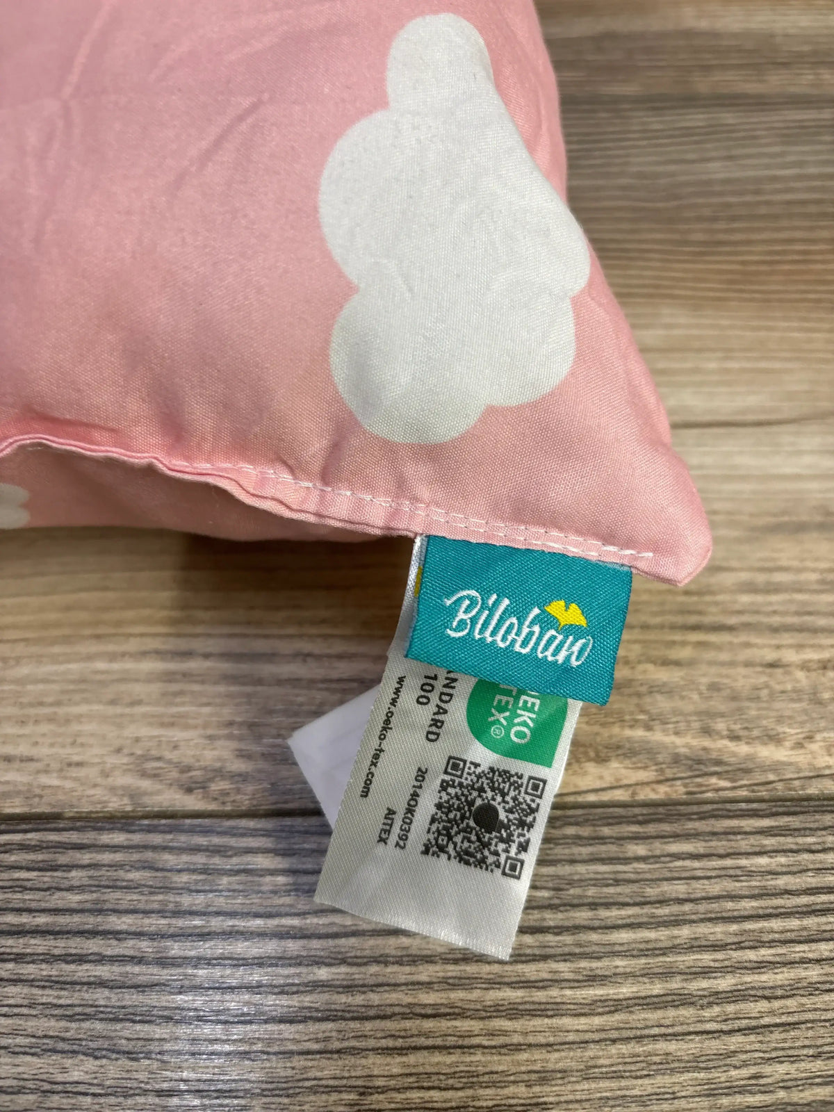 Close-up of a Biloban toddler pillow in pink with a white cloud pattern and brand tag on a wood surface