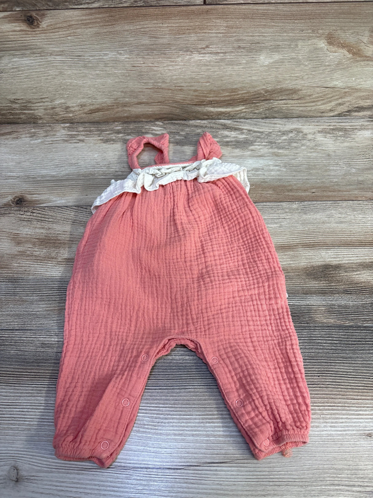 Front flat lay of pink gauze baby jumpsuit with white ruffle trim and snap closures at the legs on a wood background