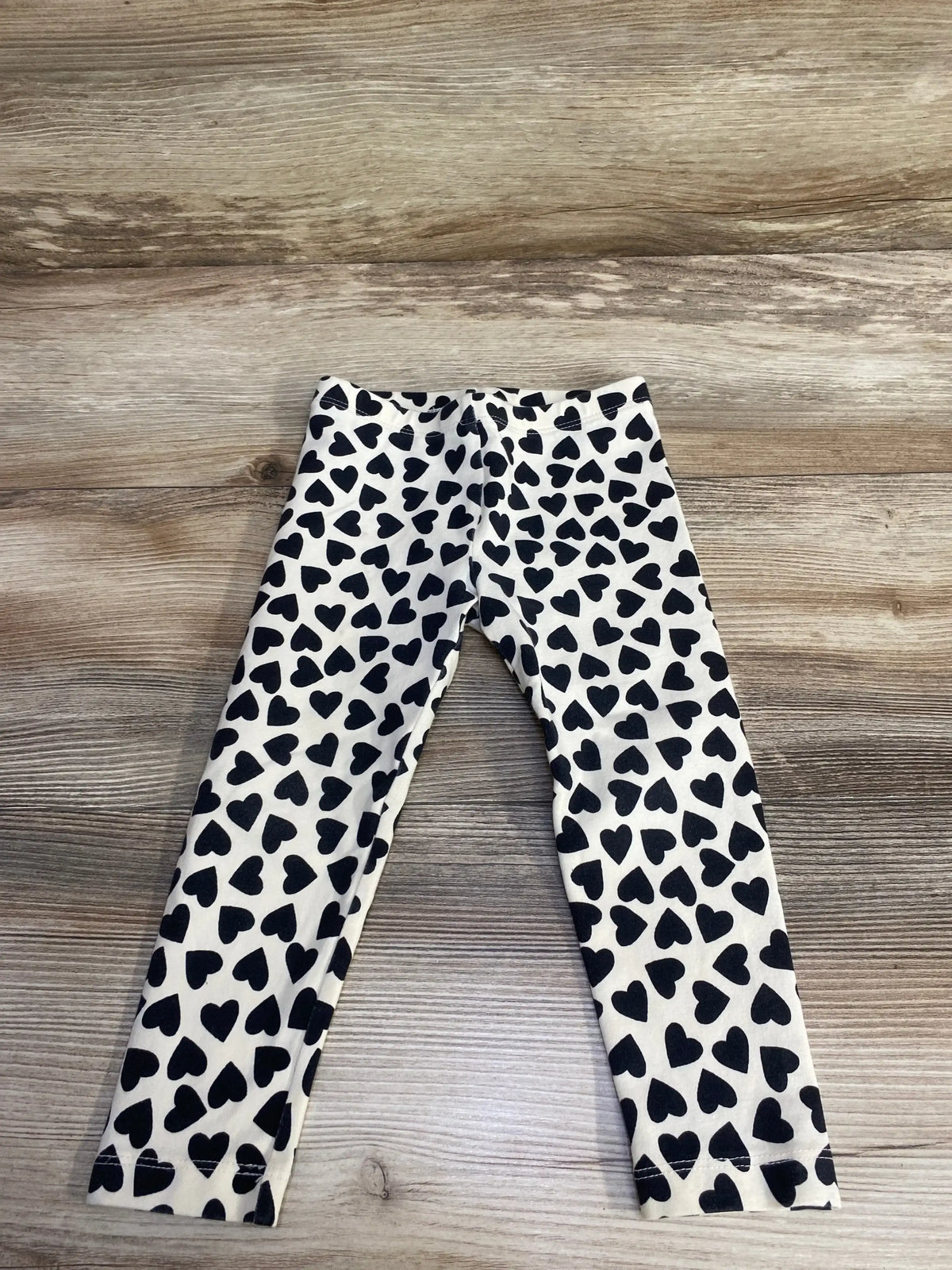 Flat lay of Tea Collection ivory leggings showcasing all-over black heart print and elastic waistband on wood plank floor