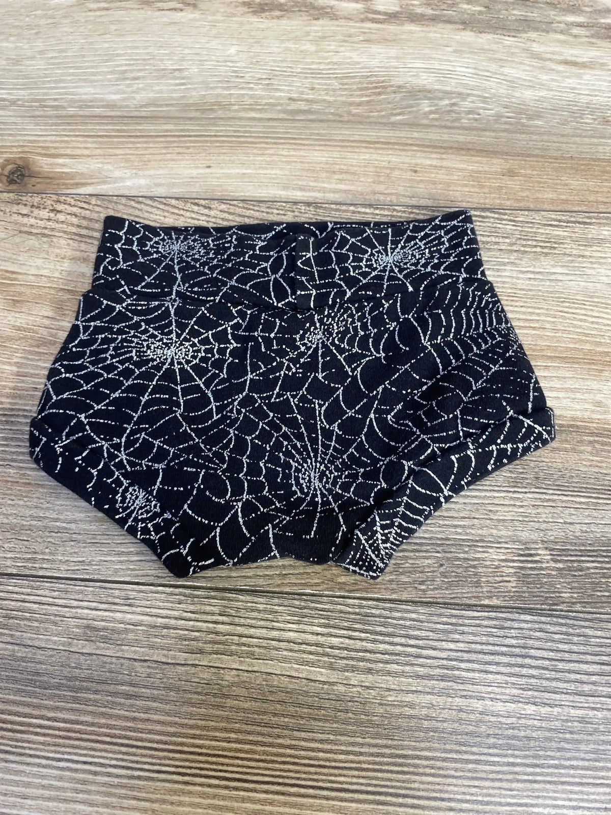 Top-down view of black baby bloomers showcasing silver spiderweb print and folded high waistband on wooden floor