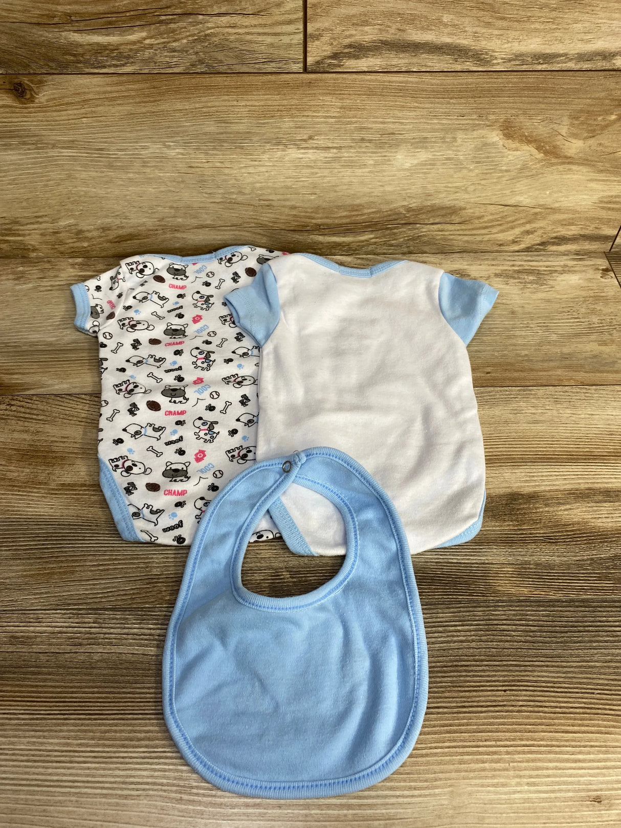 Flat lay of two baby bodysuits and a bib: one white with blue trim, one white with puppy print, plus a light blue bib on wood