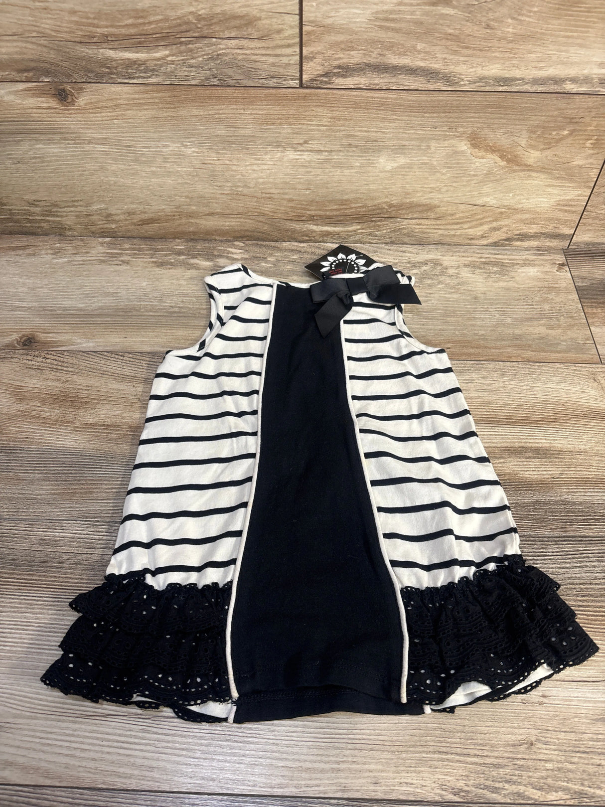 Front view of white sleeveless striped tunic in size 4T showcasing black eyelet trim and shoulder bow on a wood floor
