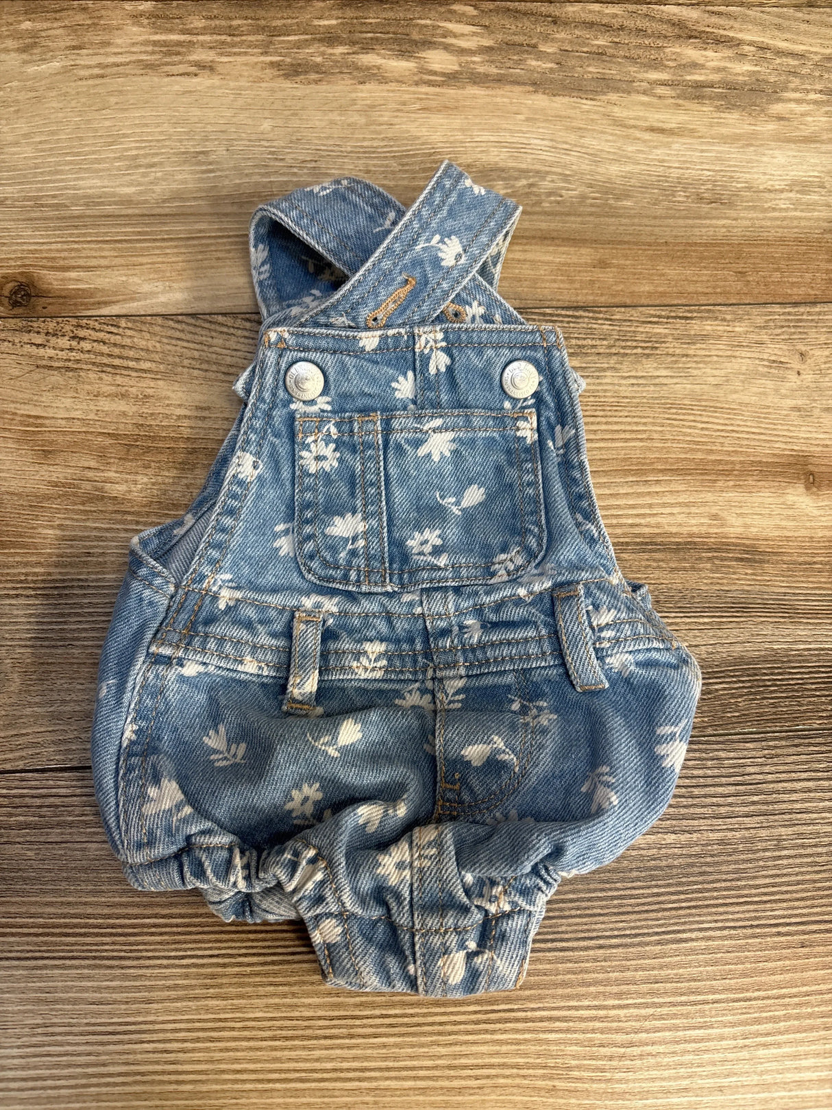 Blue floral denim baby romper with pockets and buttons on wood background