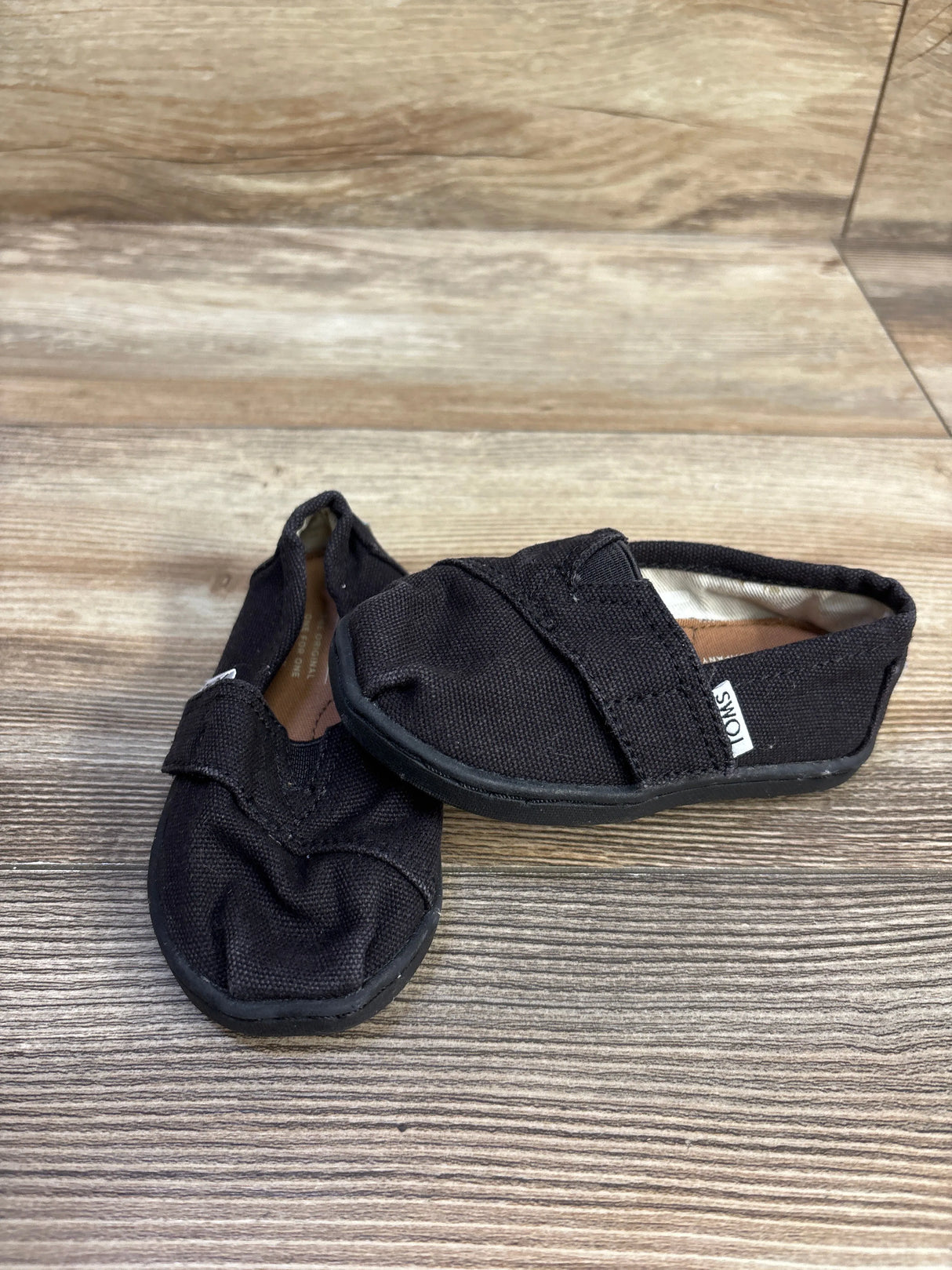 Angled view of Toms Tiny Alpargata baby shoes in black showing canvas upper, elastic V strap and rubber sole on wood background
