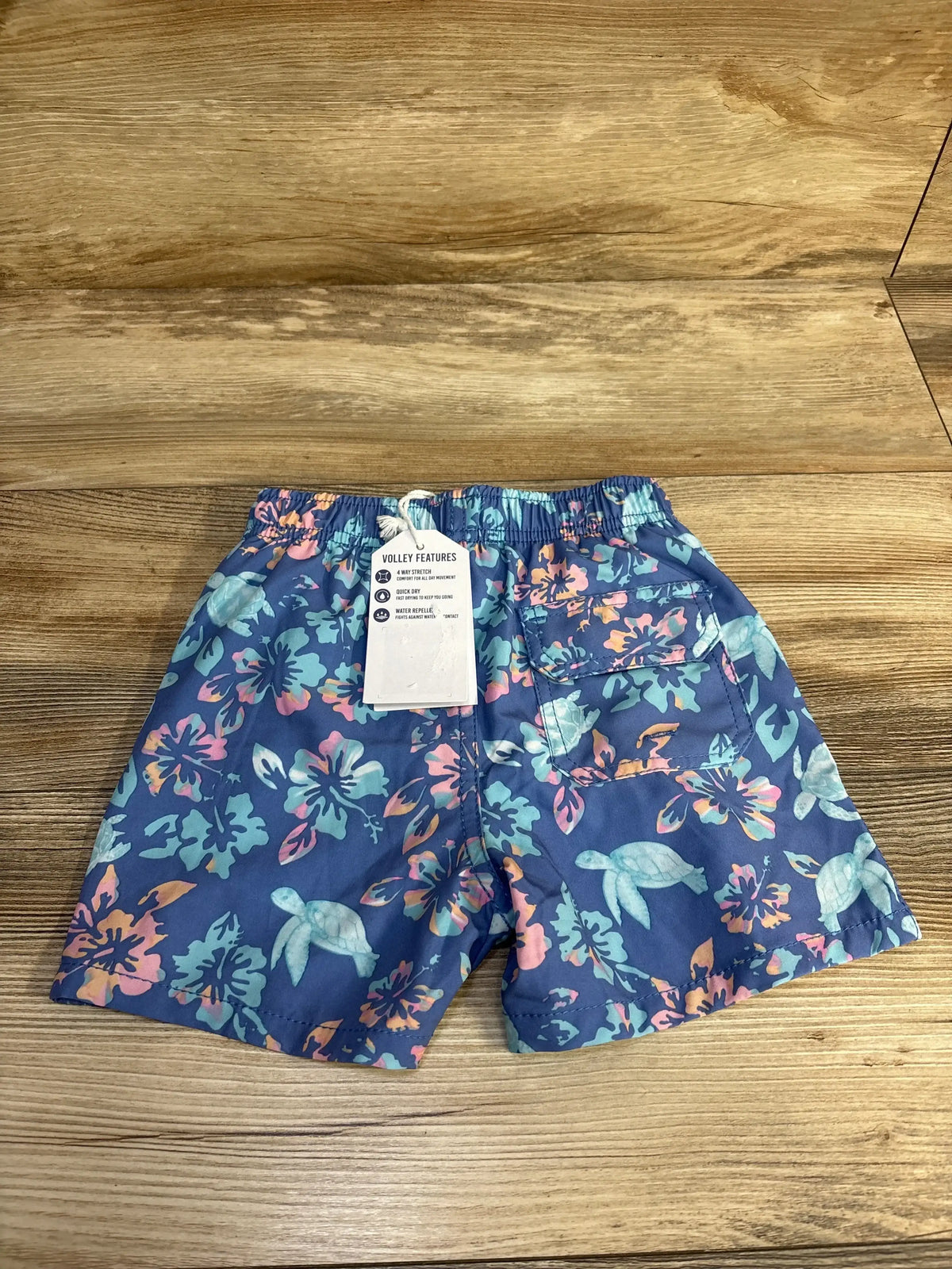 Flat-lay back view of toddler swim shorts with blue floral and sea turtle print, elastic waist and back pocket on wood