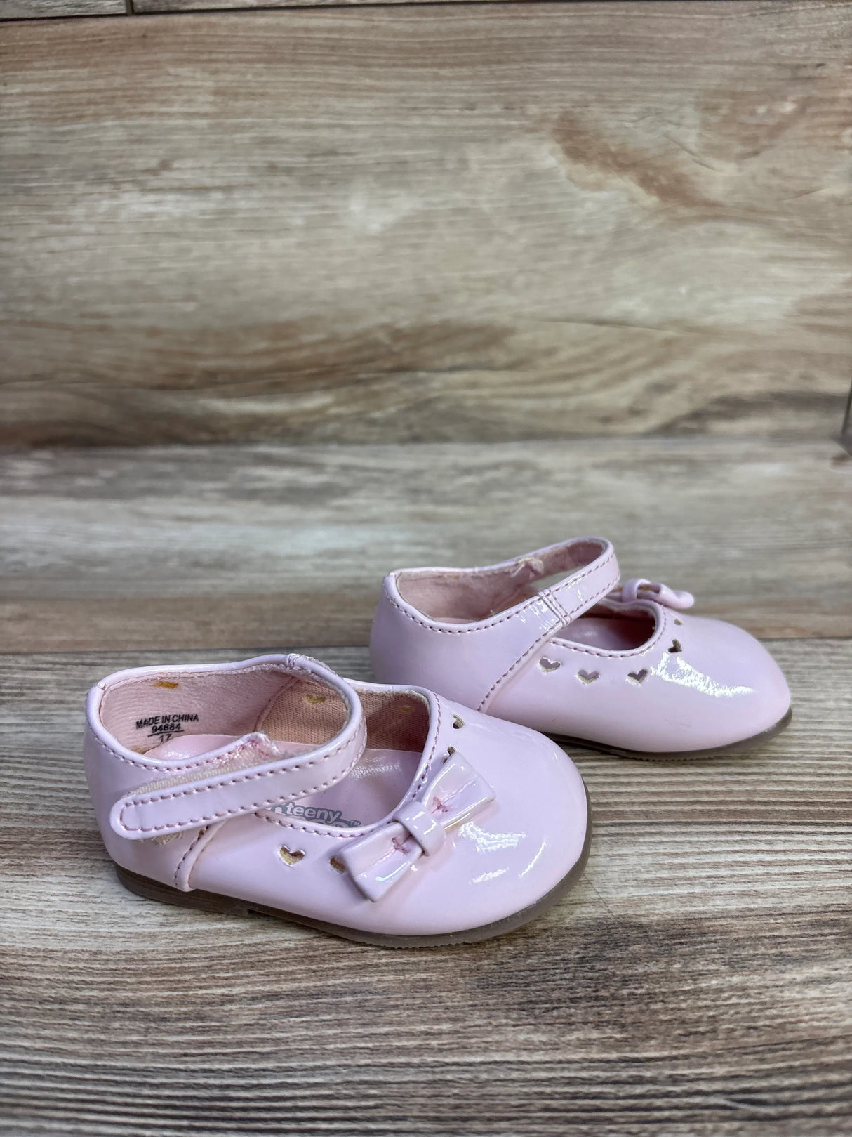 Front three-quarter view of Teeny Toes pink patent Mary Jane baby flats showing bow and heart cutout details on woodgrain background