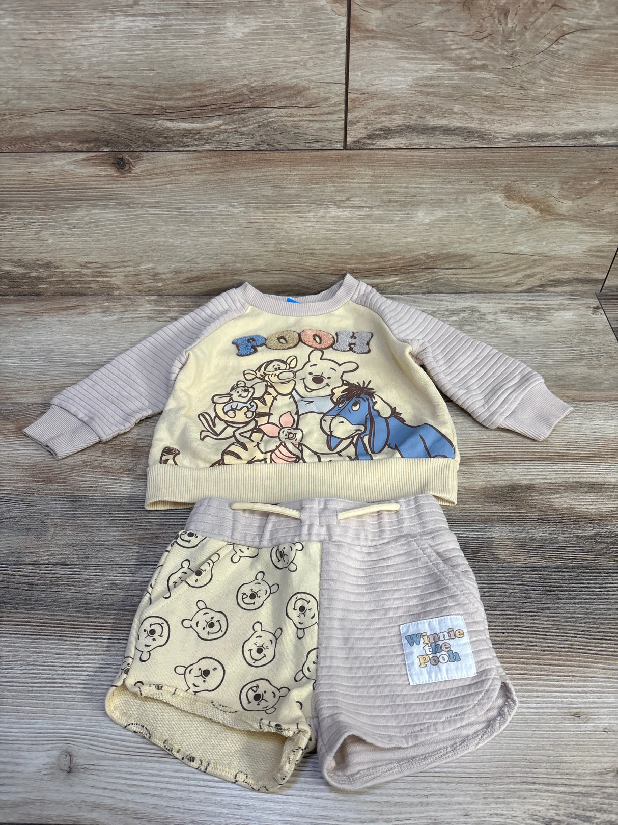 Flat lay of yellow toddler Winnie the Pooh sweatshirt and shorts set showcasing character graphic top and mixed-pattern shorts on wood background