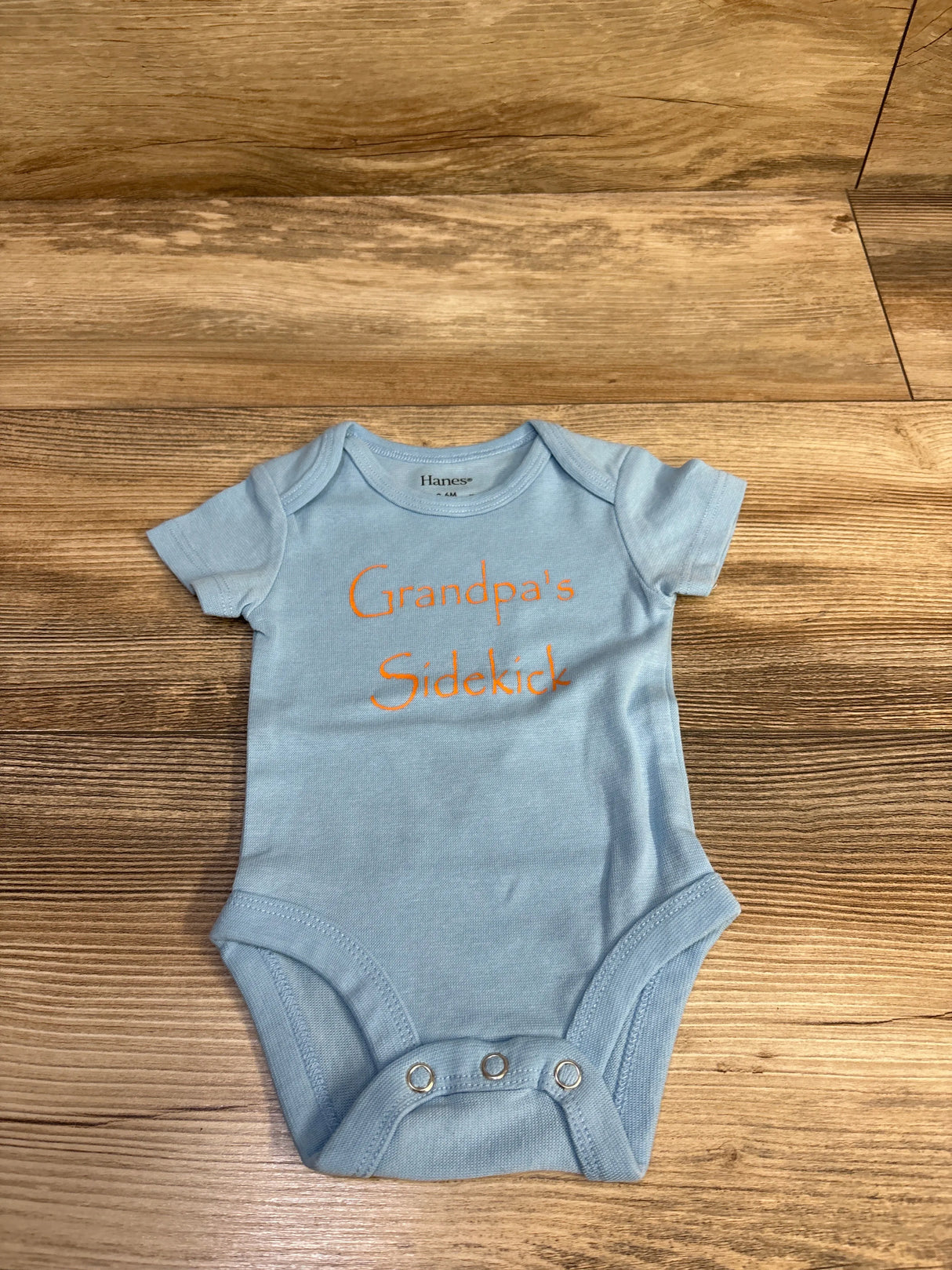 Front view of light blue Hanes baby bodysuit with orange Grandpa’s Sidekick text and three metal snaps, on wood background