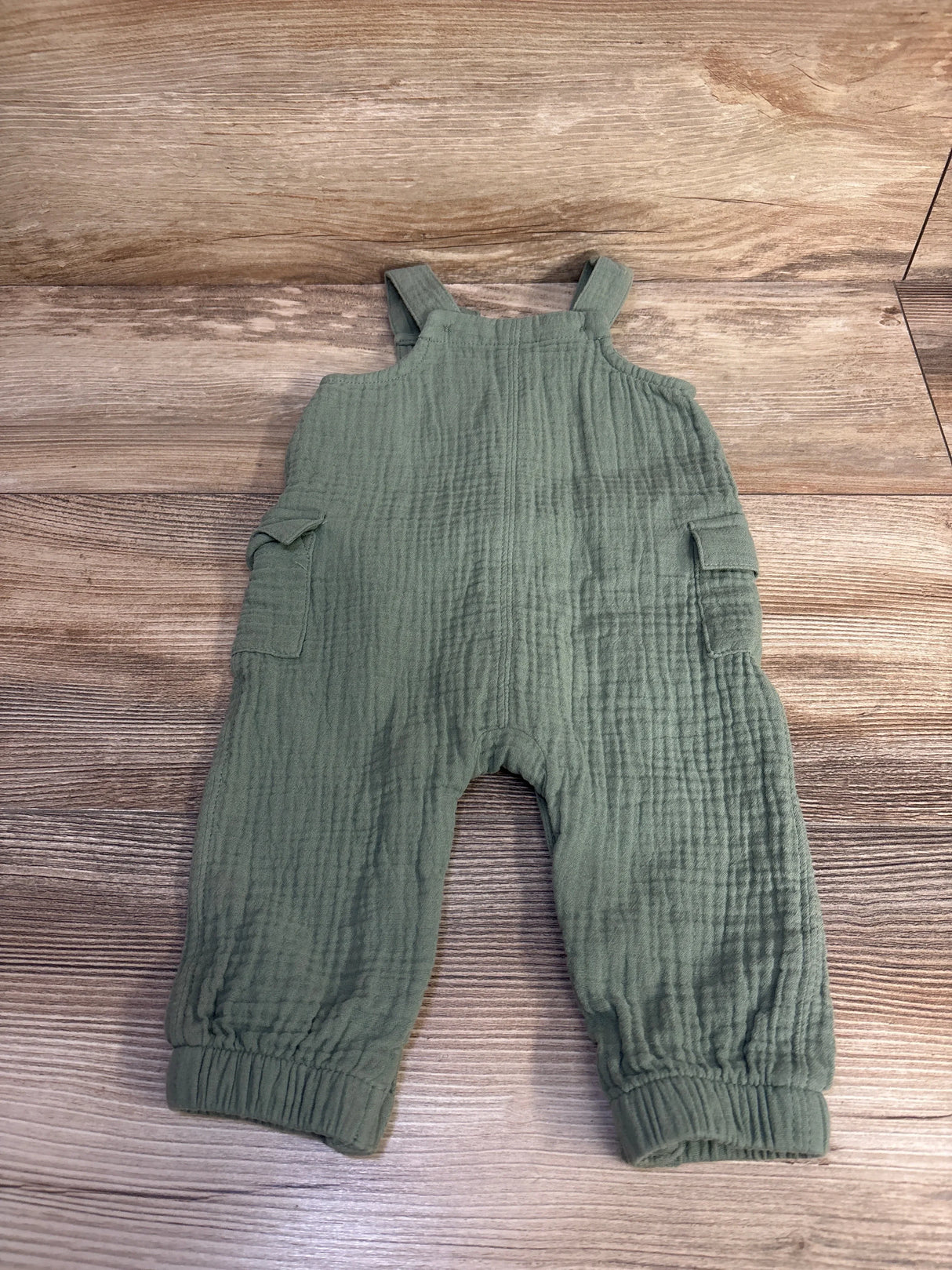 Front of Cat and Jack green gauze jumpsuit 3-6 months with textured fabric, shoulder straps, side pockets, elastic cuffs on wood