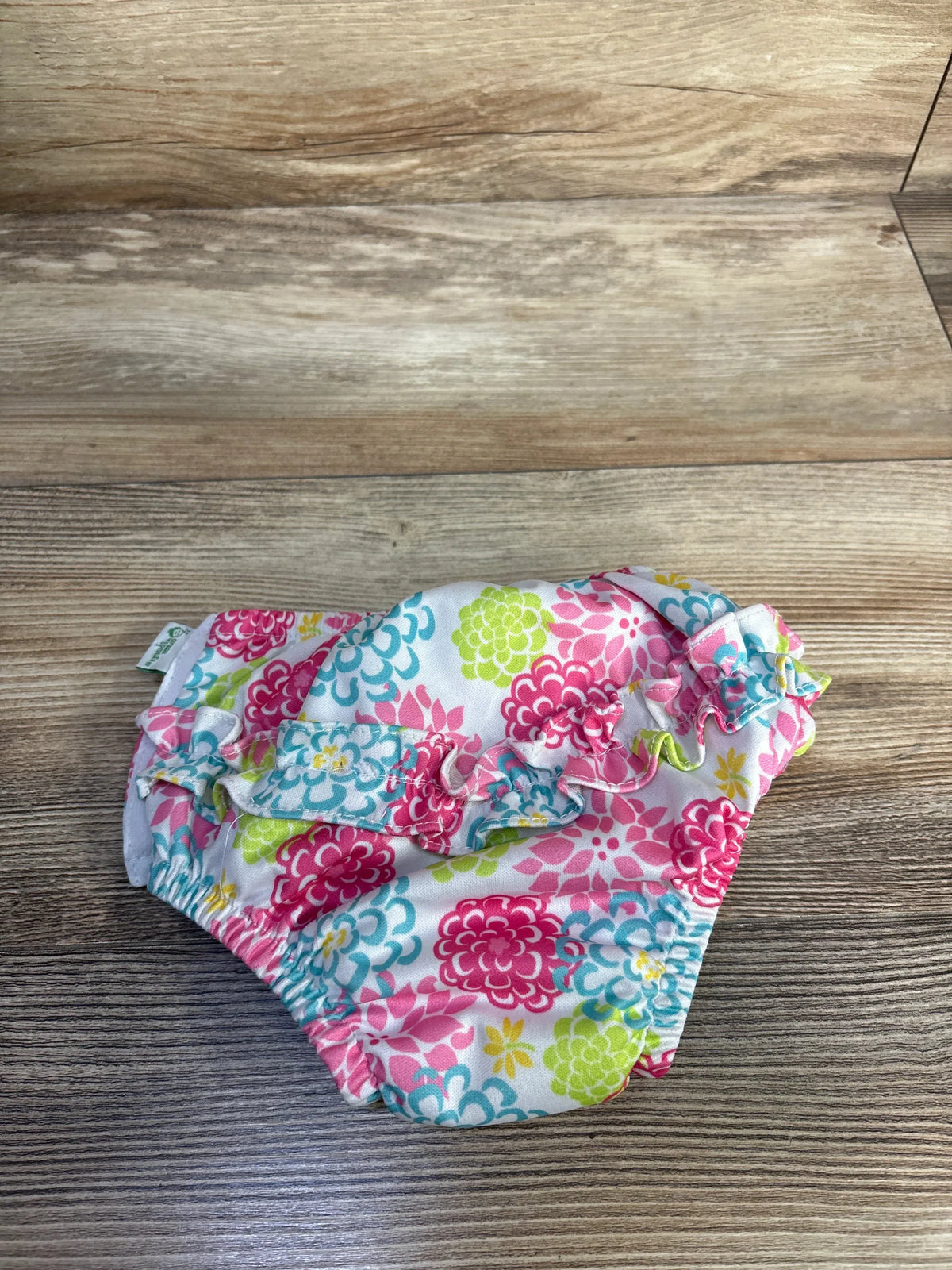 Front view of Green Sprouts floral white swim diaper showcasing ruffled leg openings on a wood grain background