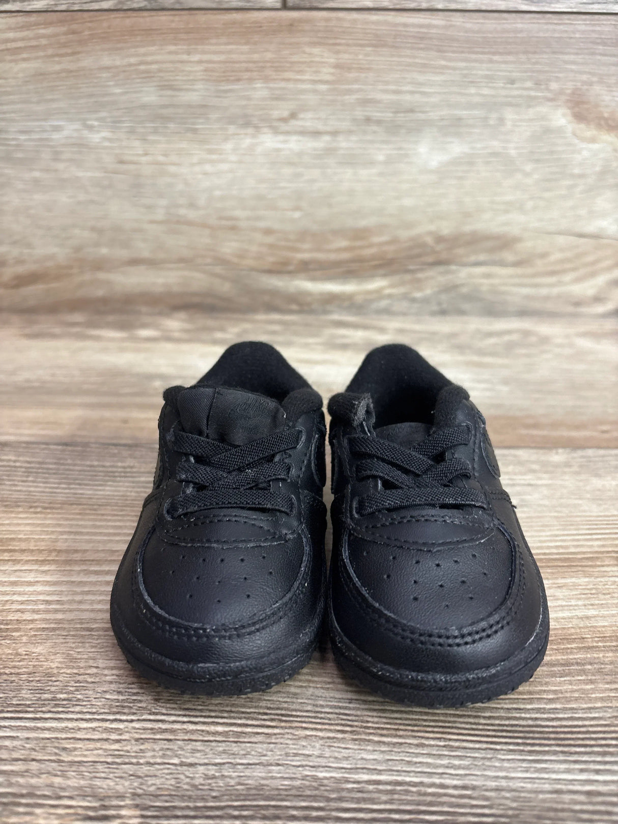 Front view of Nike Air Force 1 crib shoes showing black leather upper and laces on a wood plank background
