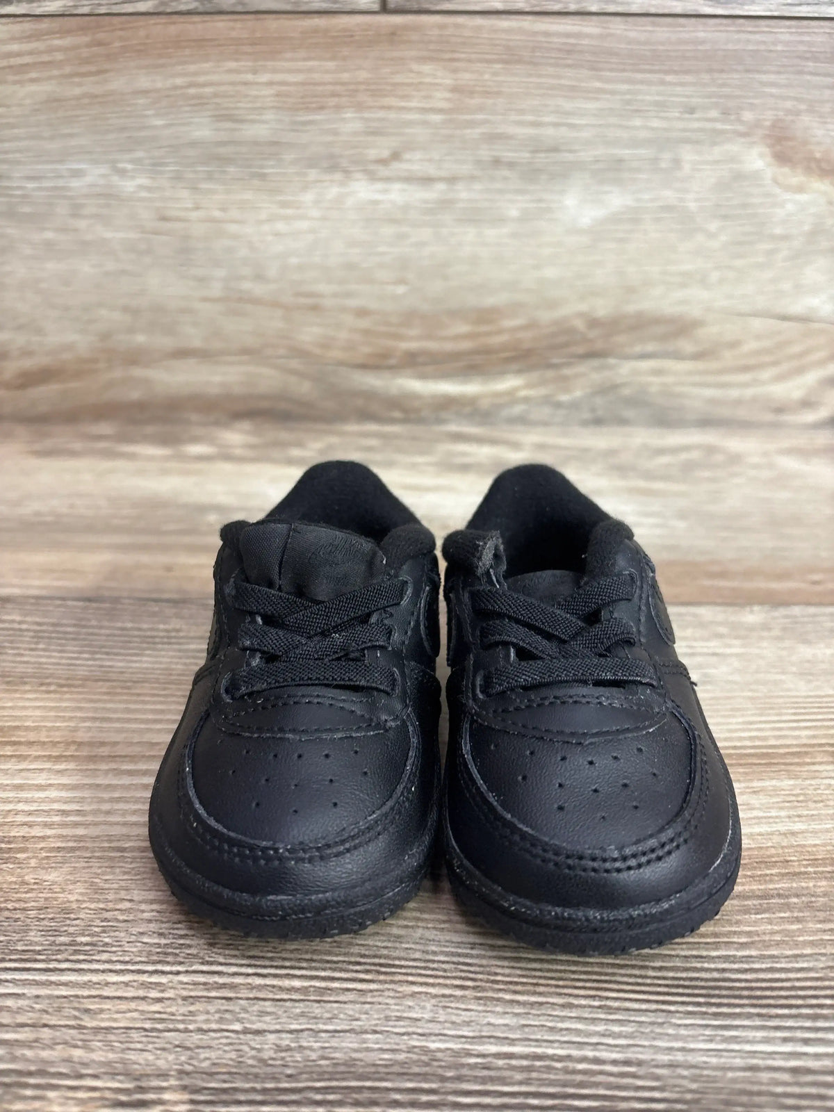 Front view of Nike Air Force 1 crib shoes showing black leather upper and laces on a wood plank background