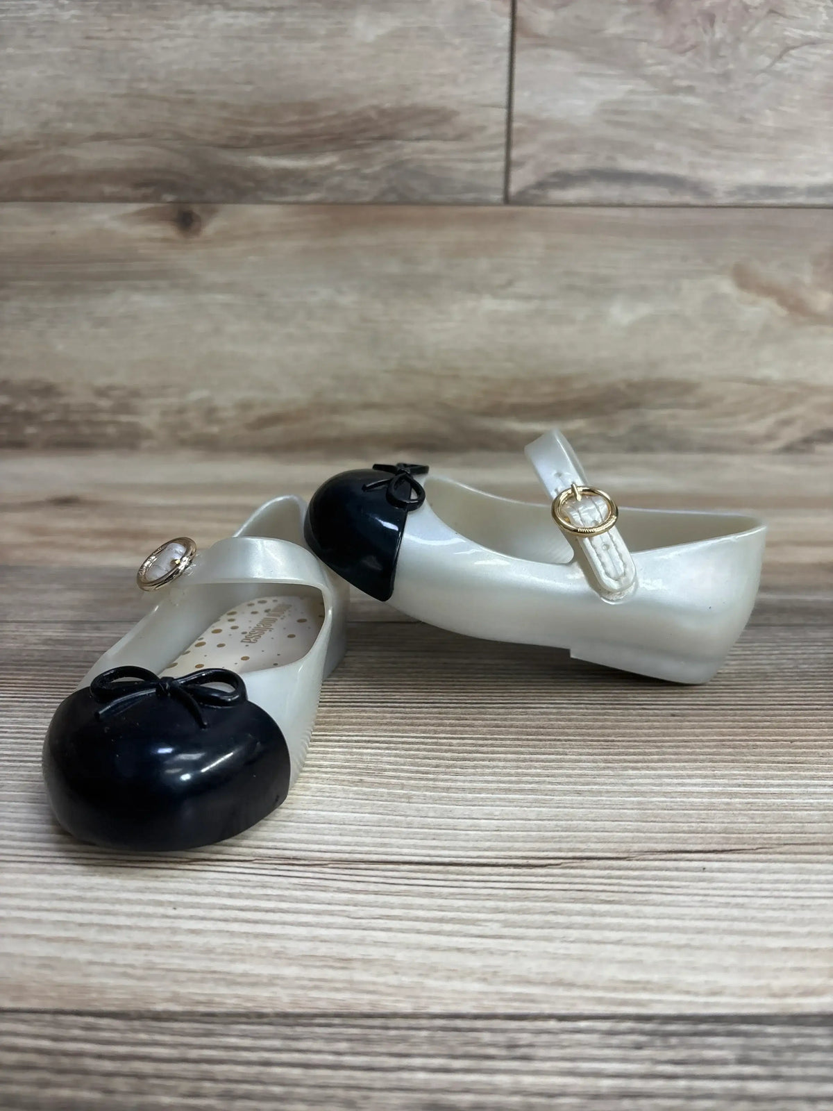 Pair of children's black and white Mary Jane shoes with gold buckles on wooden surface
