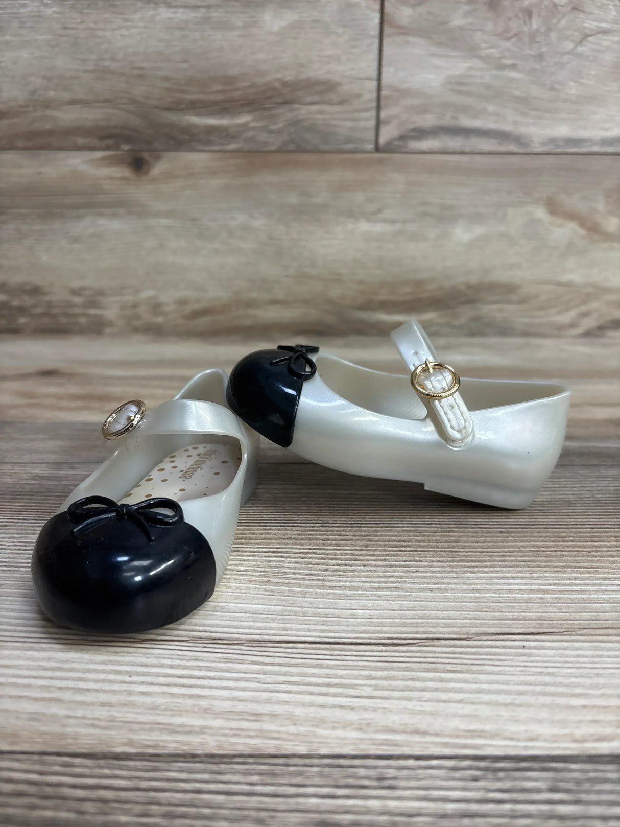 Pair of children's black and white Mary Jane shoes with gold buckles on wooden surface