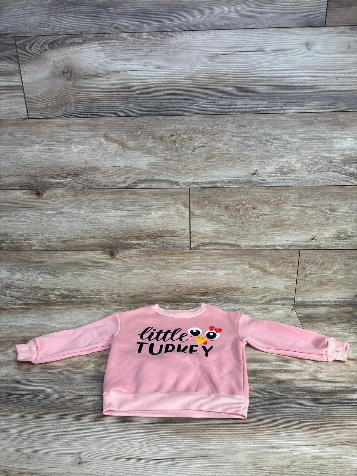 Pink toddler sweatshirt with 'little TURKEY' text and turkey graphic, laid on wood floor