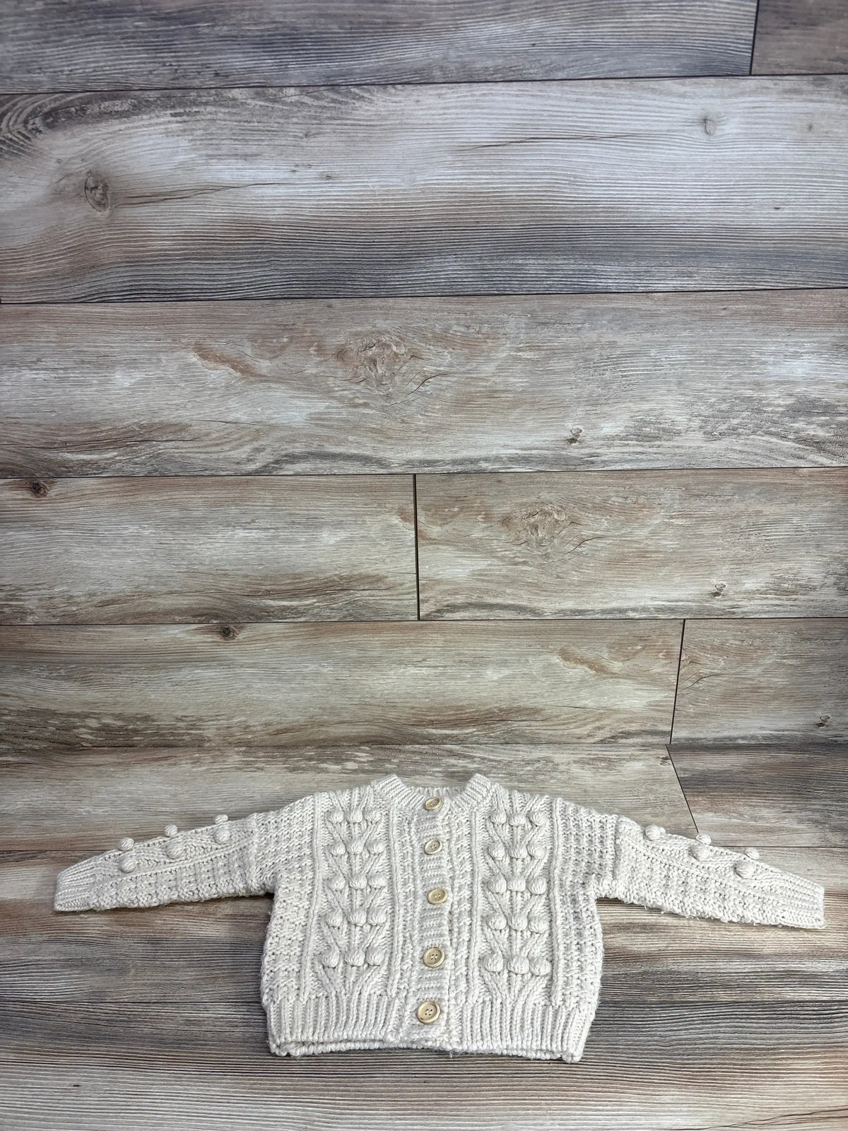 Cream knit baby cardigan sweater with wooden buttons, textured design, laid on wood floor