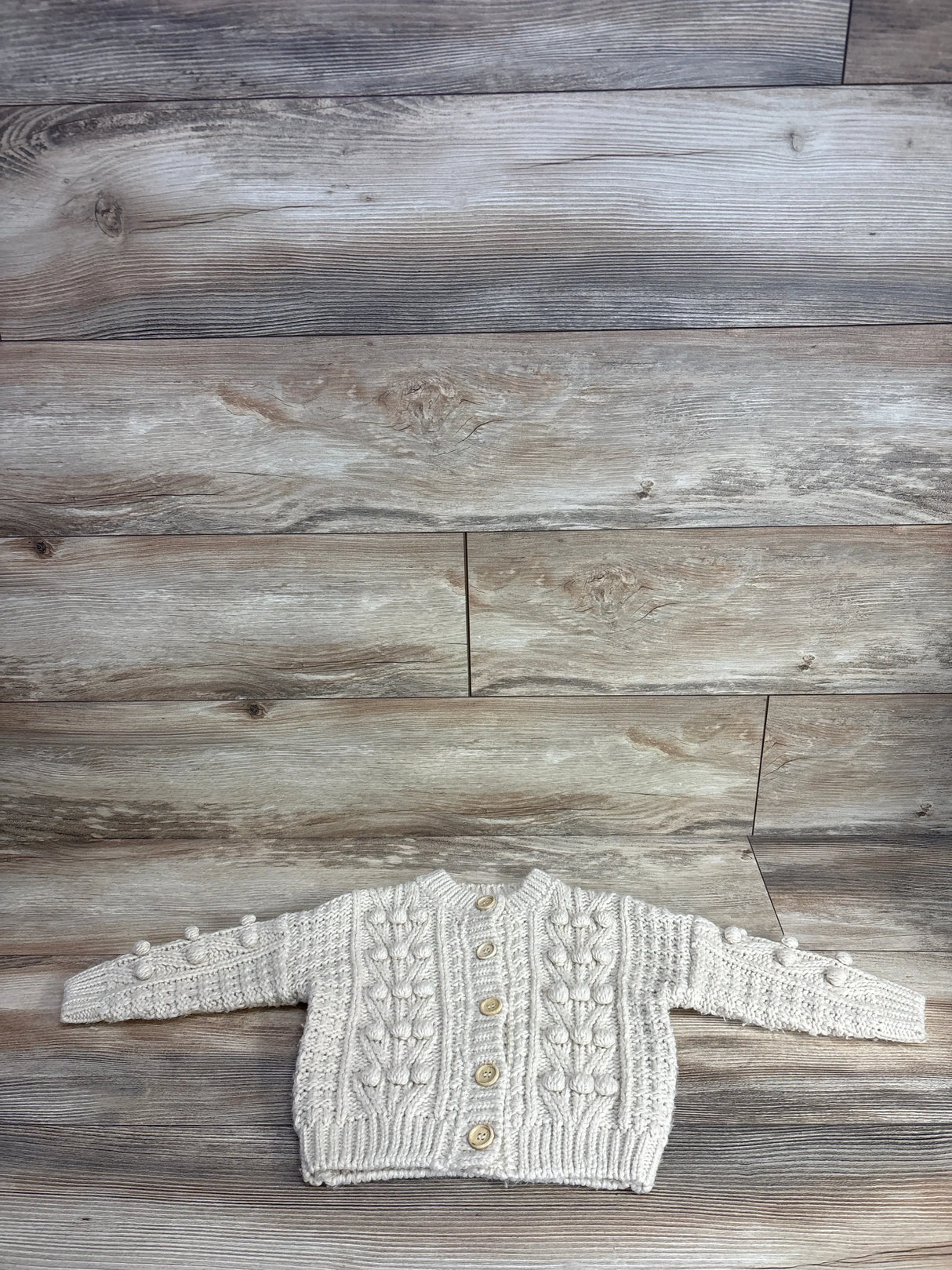 Cream knit baby cardigan sweater with wooden buttons, textured design, laid on wood floor