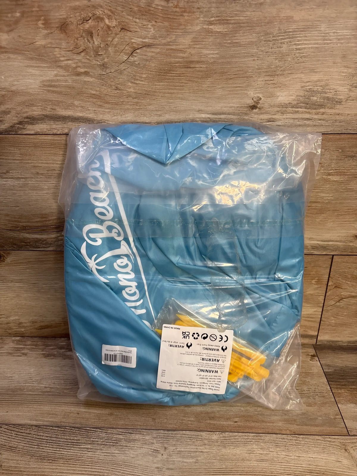 Top-down view of folded blue Monobeach pop-up baby sun tent in clear plastic packaging with yellow stakes on wood floor