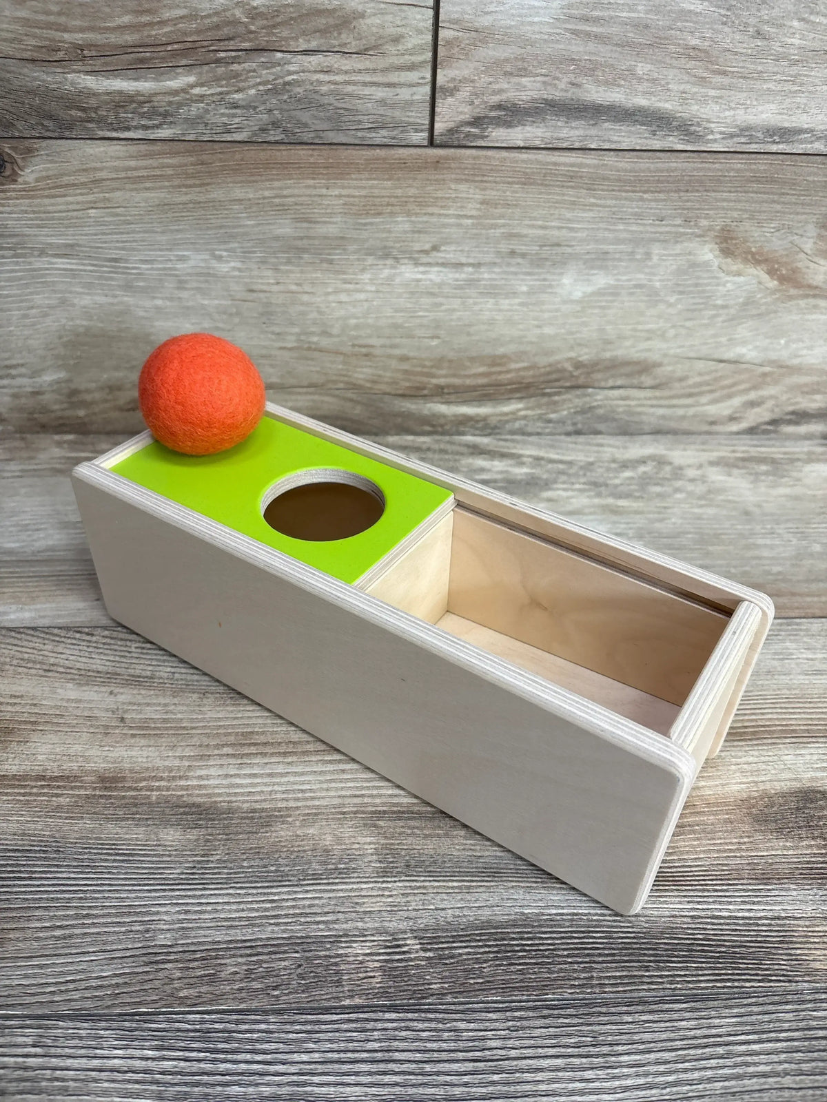 Angled view of Lovevery Sliding Top Box showing green sliding top with round hole and orange felt ball on wood-grain surface