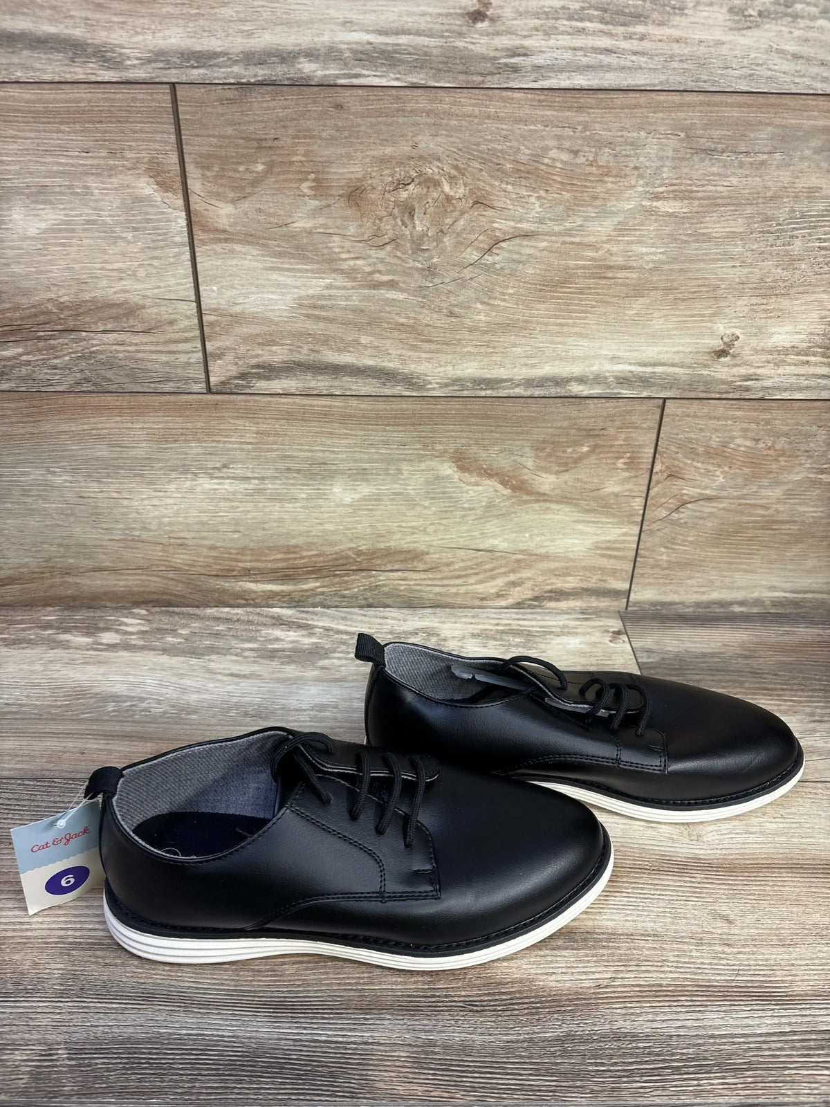 Three-quarter view of Cat & Jack Alfi dress shoes showcasing black lace-up uppers and white layered soles on a wood-patterned floor
