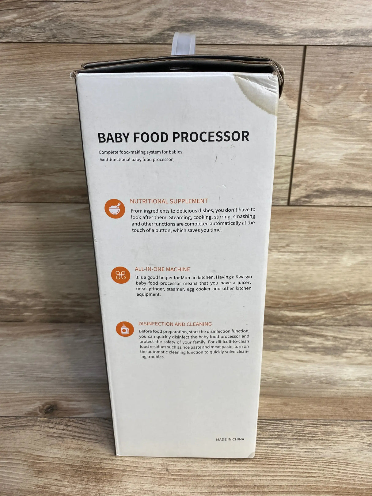 Front view of KWASYO baby food maker box showing BABY FOOD PROCESSOR headline and icons, dented corner, wooden floor