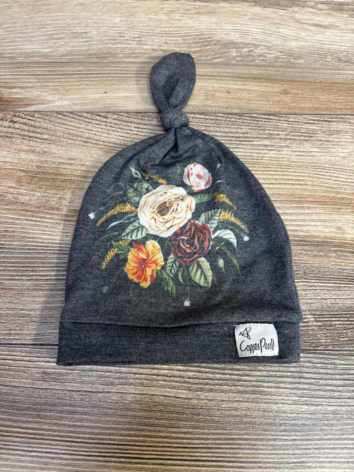 Top view of gray Copper Pearl baby top-knot hat showcasing multicolor floral print and branded label on wood tabletop