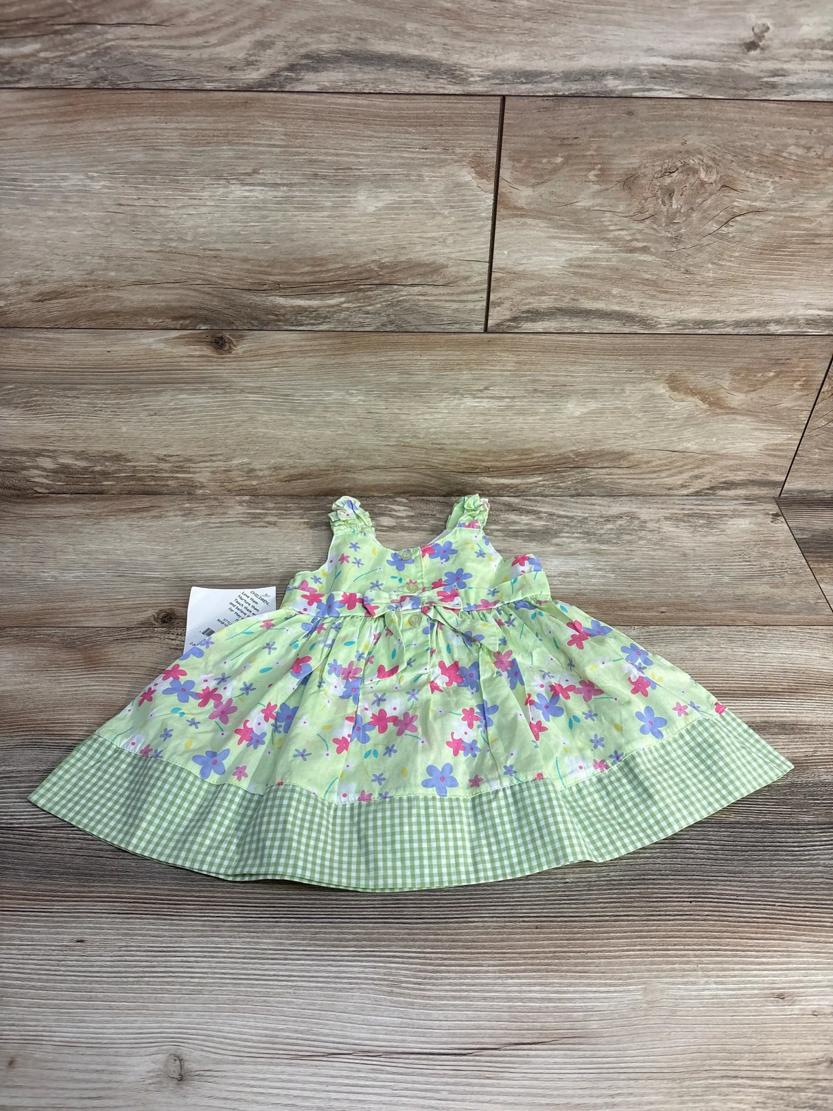 Flat lay of green baby dress with pink and purple floral print, ruffle shoulder straps, bow at waist and green gingham hem on wood background