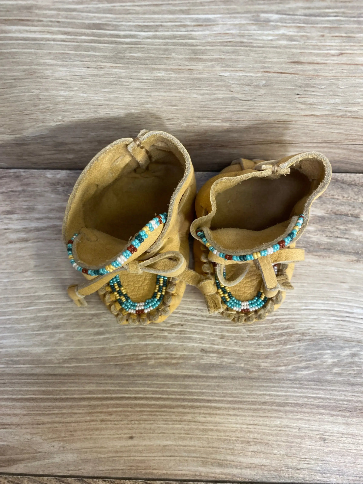 Top view of yellow suede baby moccasins size 2C showing turquoise and brown beadwork, fringe, and tied laces on wood