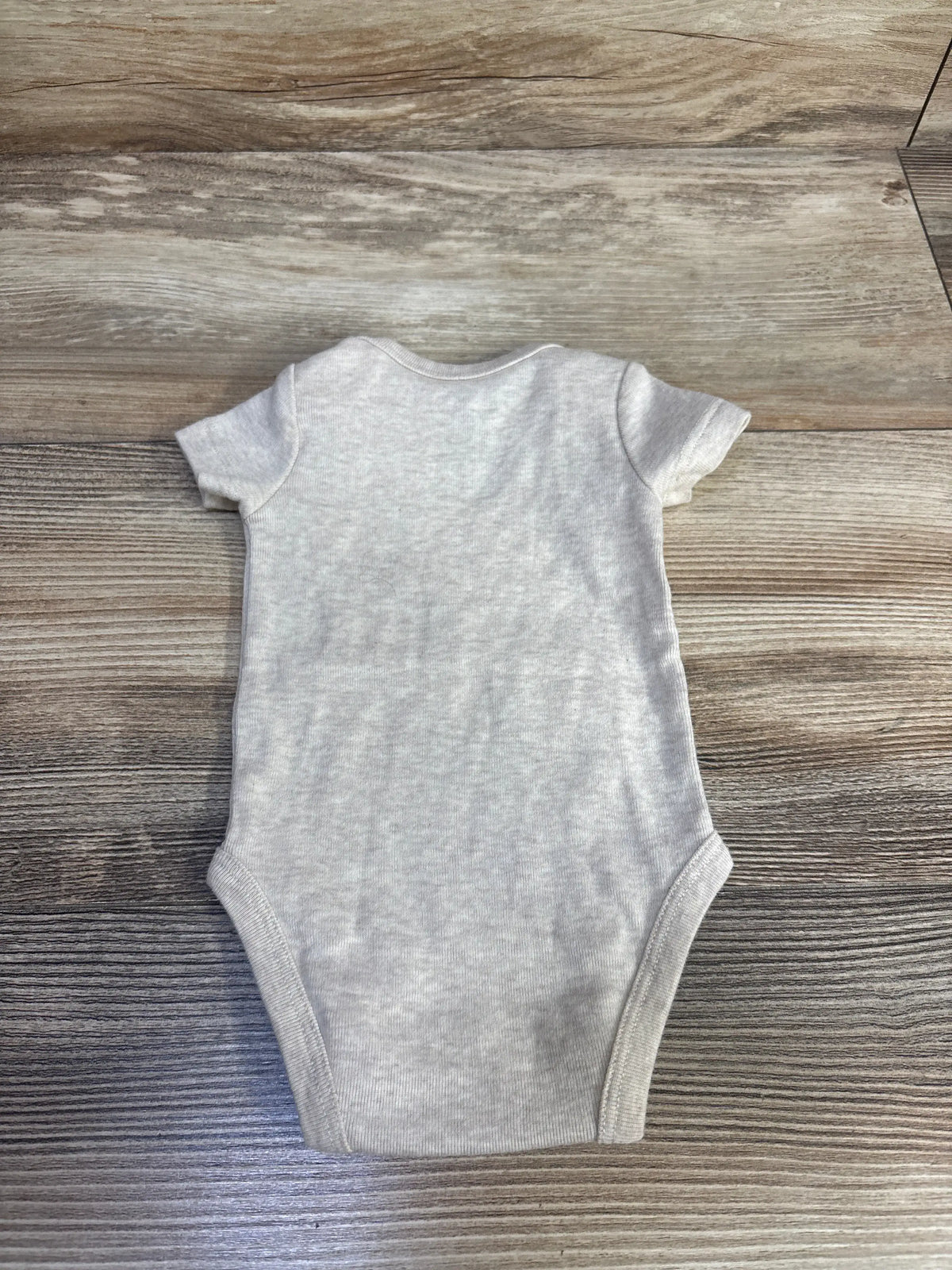 Front view of Nike beige newborn bodysuit showcasing short sleeves and rounded neckline with wood-grain background